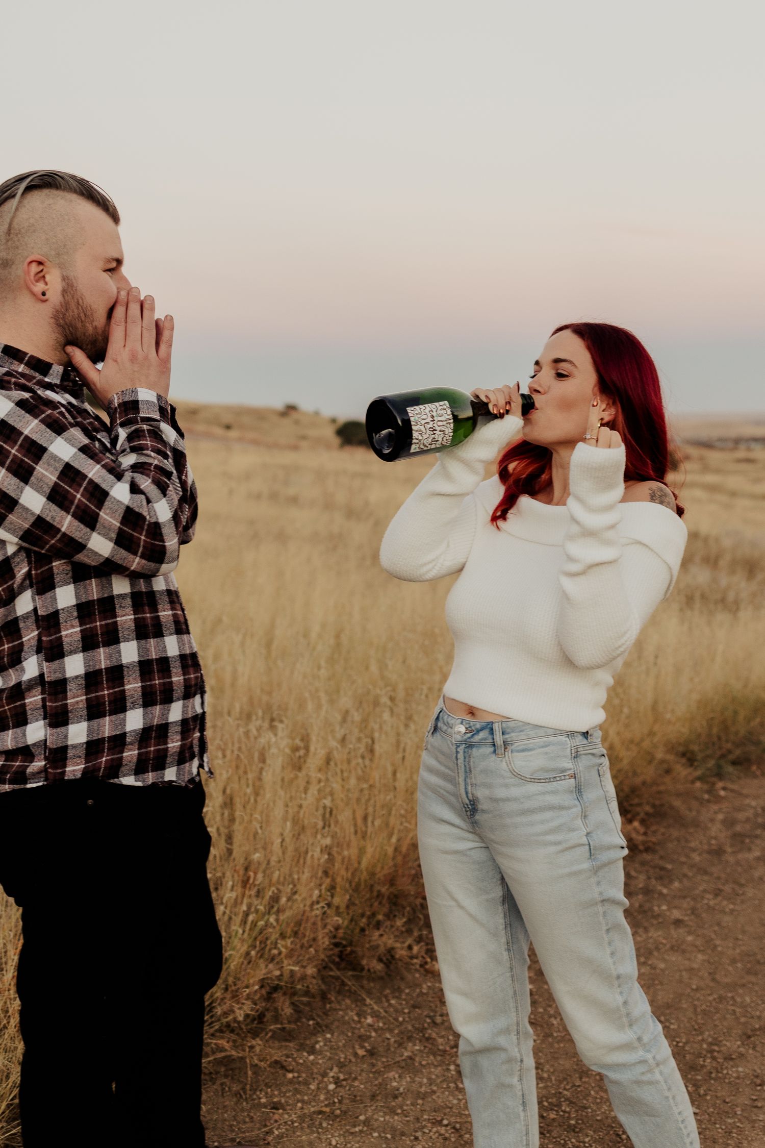 Two people share drinks while standing in a golden field at sunset.