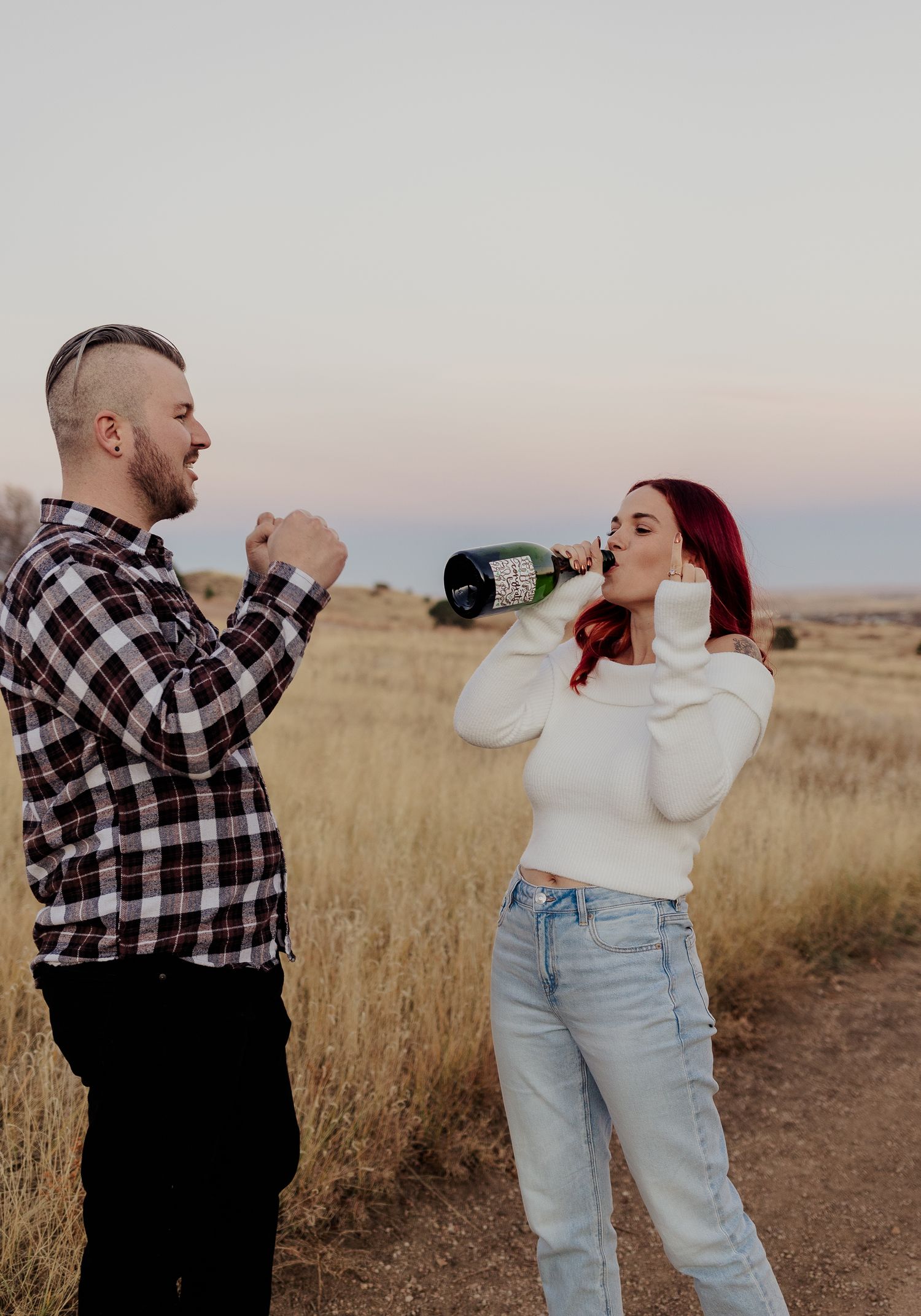Two people playfully share white beverages while standing in a golden field at sunset.