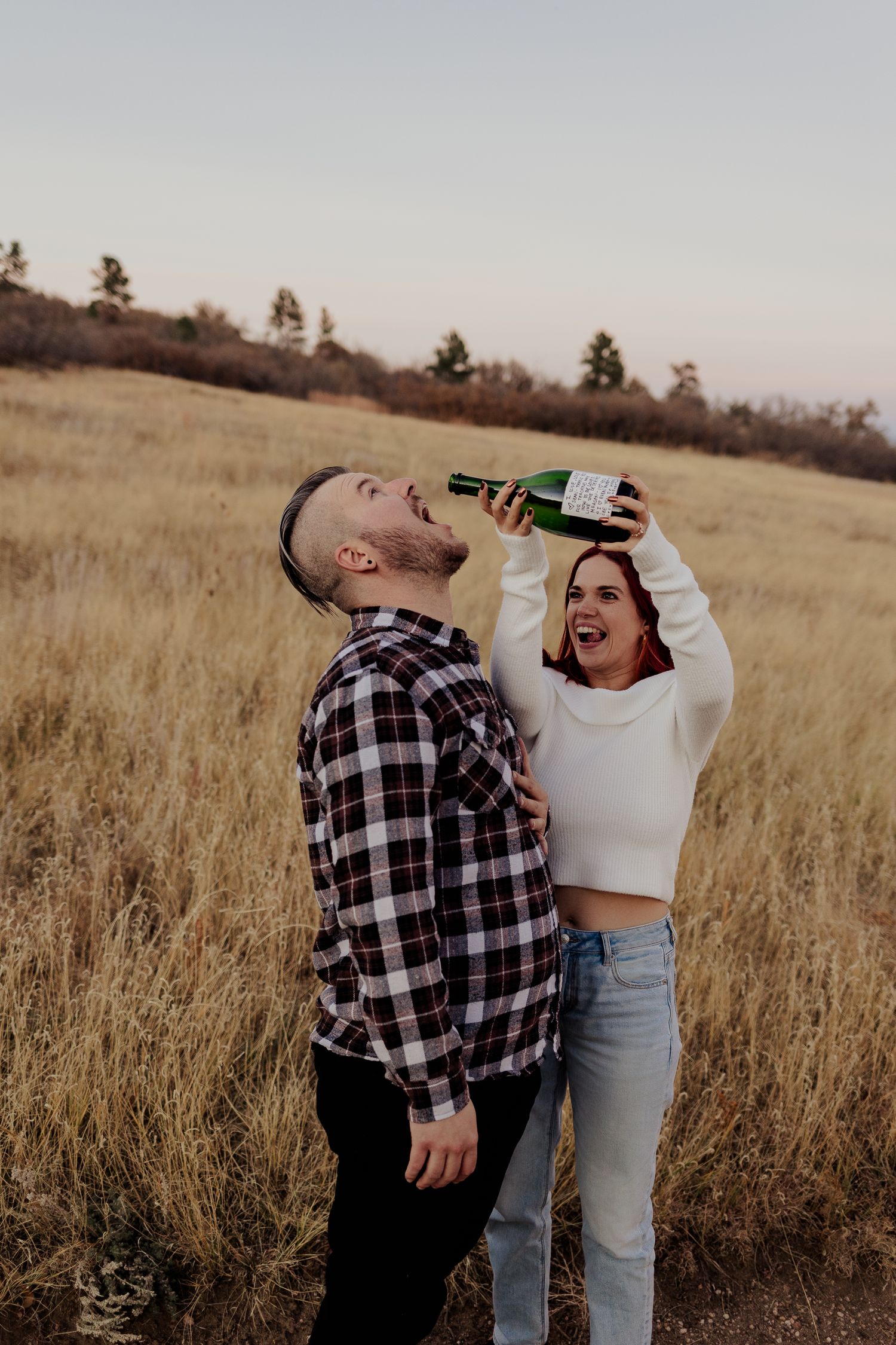 Two people in casual attire share drinks and laugh together while standing in a golden wheat field at sunset.