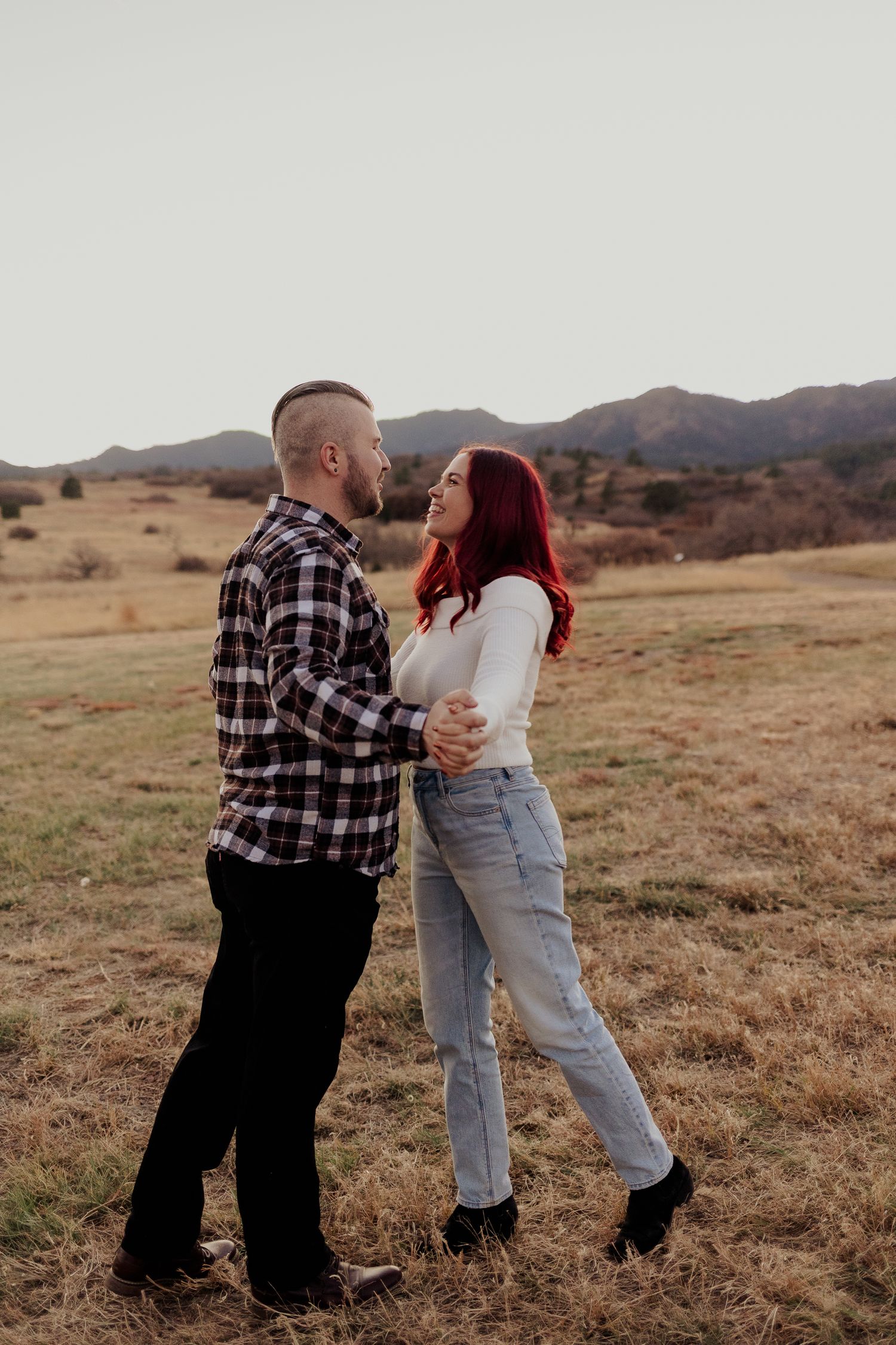 A couple in plaid and jeans dance together in a scenic golden field with mountains in the background during sunset.