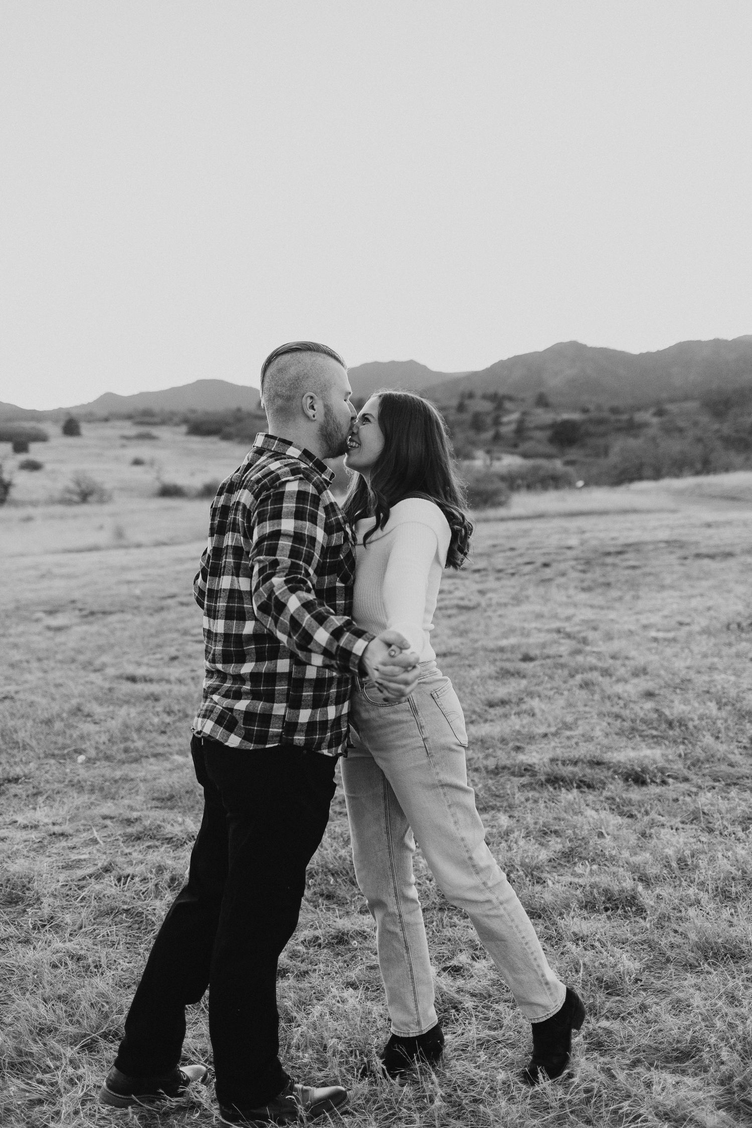 Two people in casual attire playfully interact in a scenic rural field with mountains visible in the background.