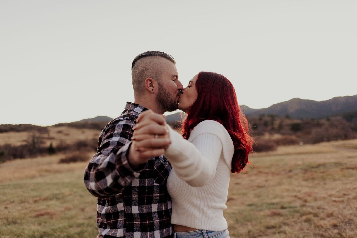 A couple in rustic outdoor attire embraces in a scenic field at sunset with mountains in the background.