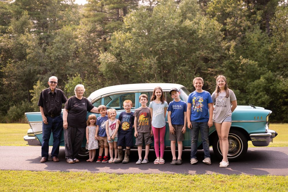Large group gathering around a classic turquoise Chevrolet for photos.