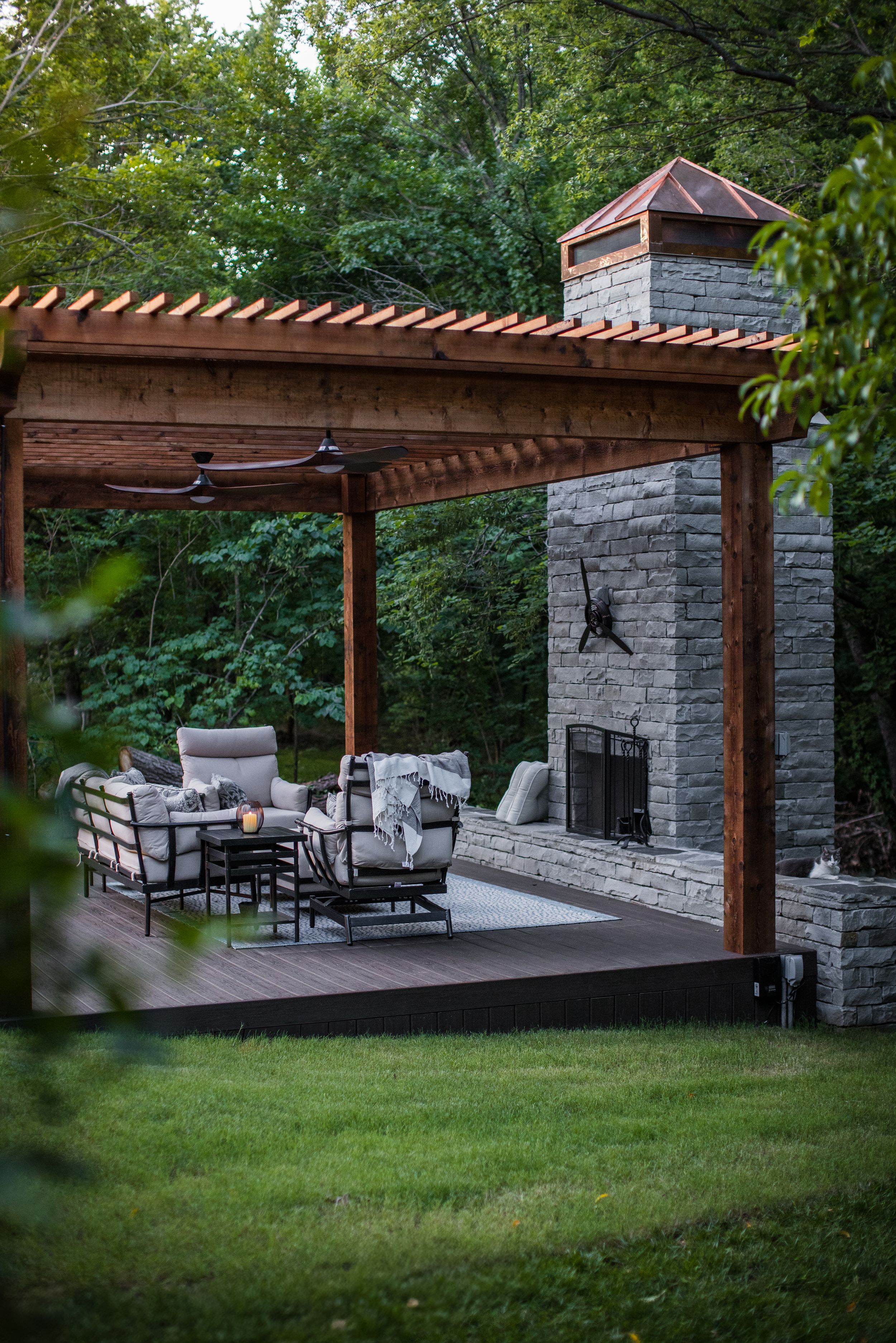 Cozy outdoor patio space with wooden pergola, stone fireplace and comfortable seating arrangement nestled in a green backyard.