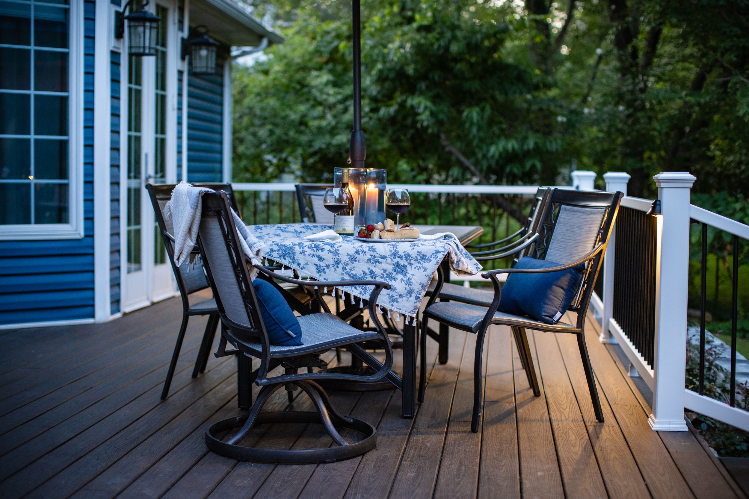 Cozy outdoor deck setting with patio furniture, blue cushions, and candlelight at dusk beside a blue house.