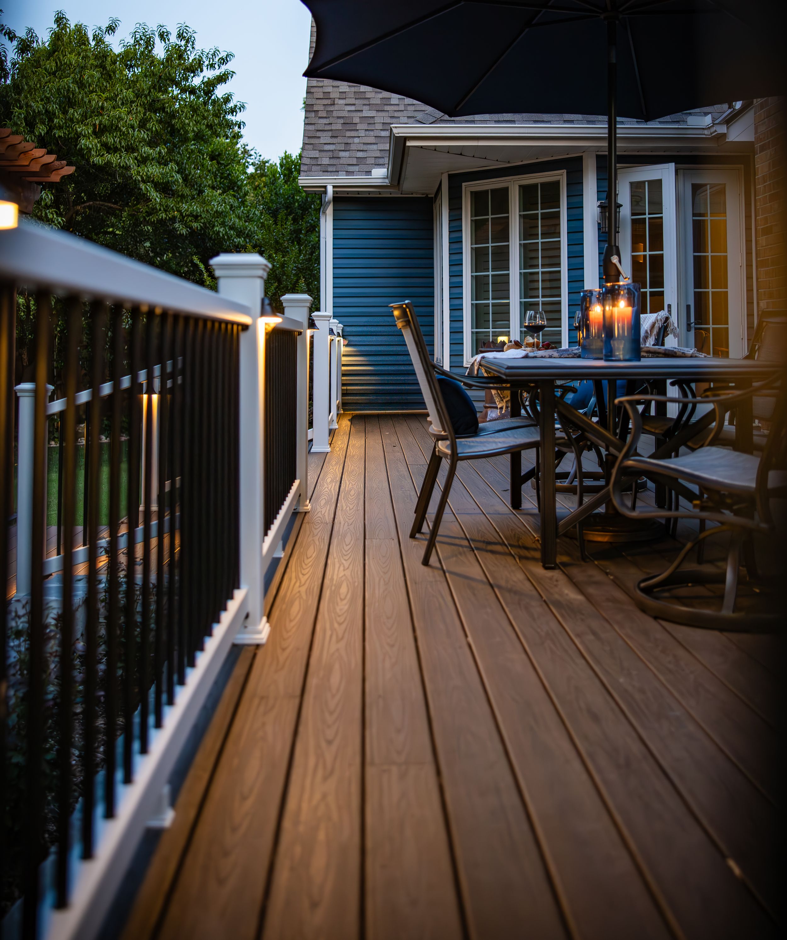 Cozy deck with string lights features wooden railings, outdoor furniture and a blue house exterior at dusk.