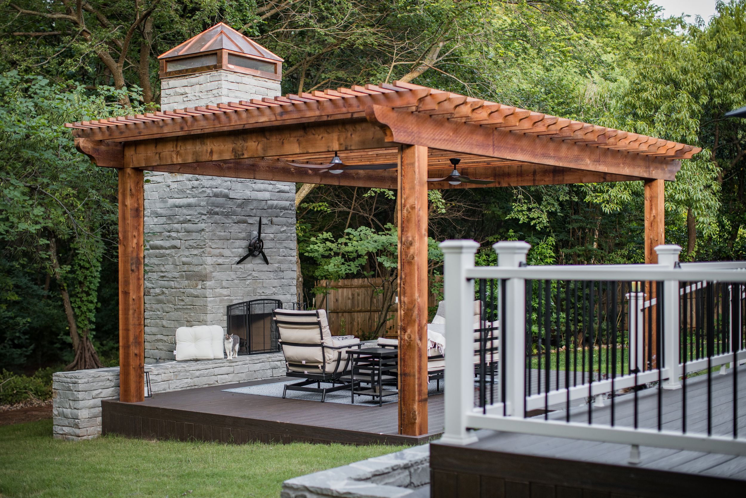 A modern outdoor pergola with cedar beams features a stone accent wall, comfortable seating, and black metal railings.