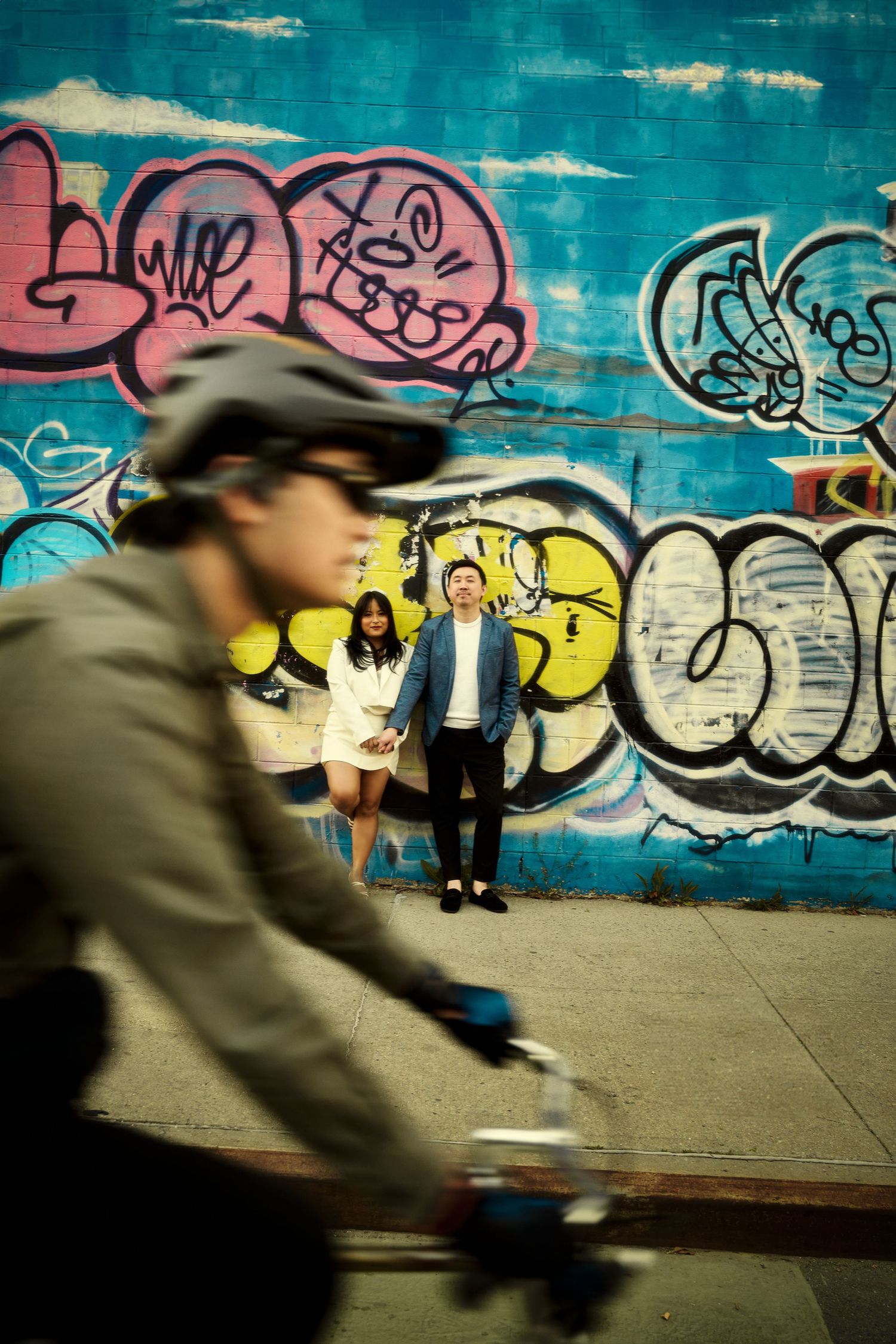A cyclist passes by a colorful graffiti wall while people pose against the street art background with an urban vibe. Engagement  in Williams