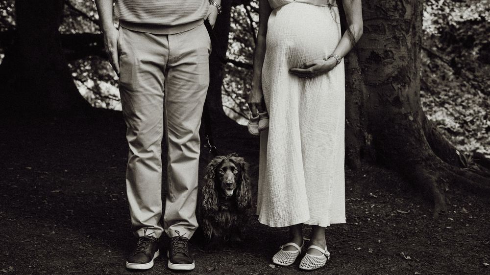 Expectant couple with their dog during an outdoor maternity photoshoot, captured in black and white.