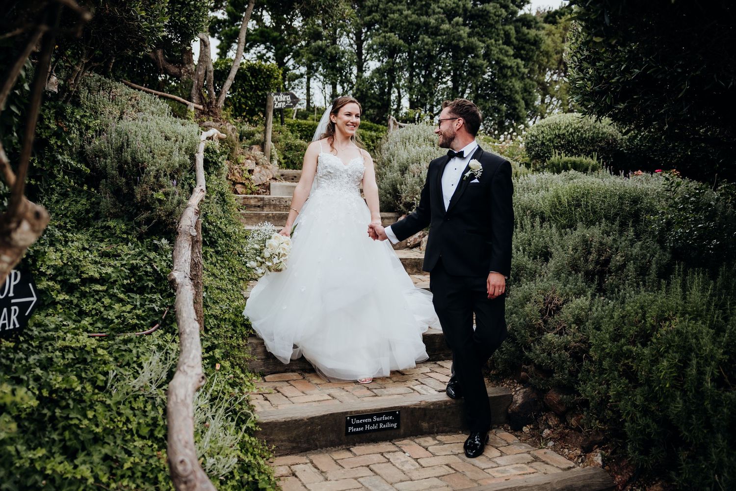 Wedding guests throw confetti as a couple walks down the aisle under natural outdoor lighting.