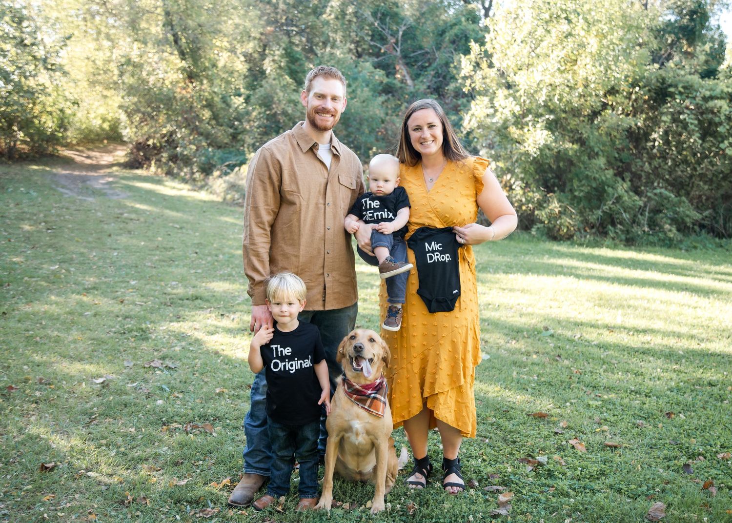 A family stands on grass with their brown dog wearing matching black and white t-shirts in an outdoor setting.