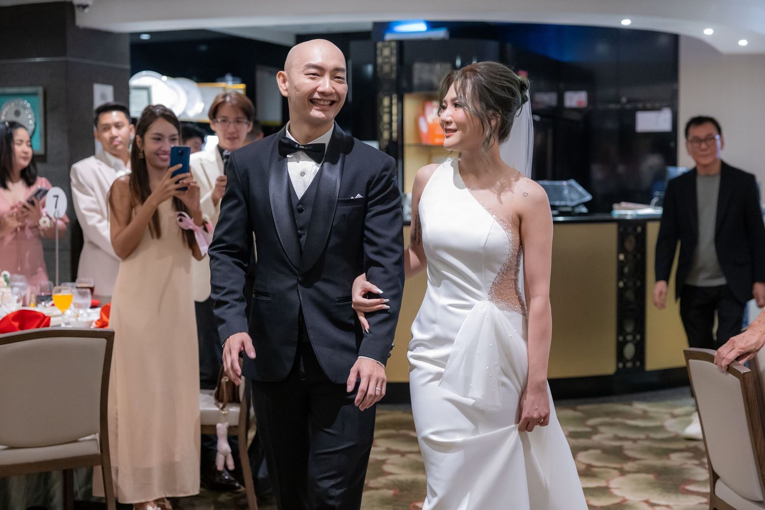 Wedding guests raise their glasses in celebration at an intimate reception in front of traditional Chinese decorations.