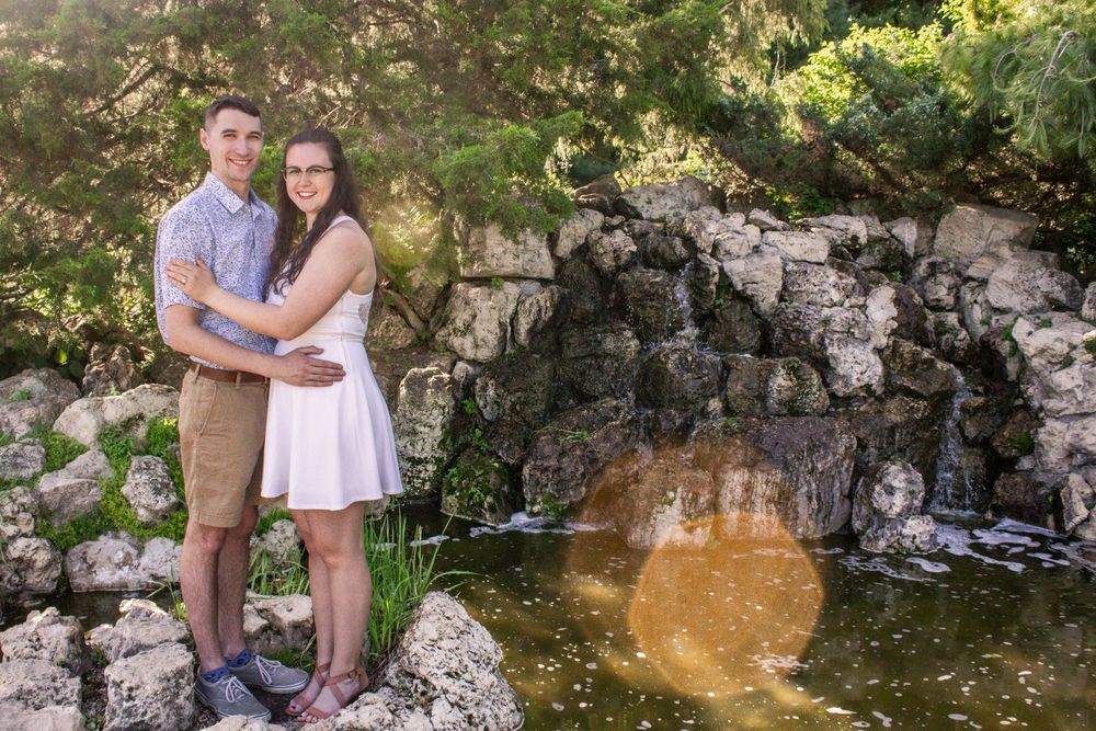 A romantic couple poses together near a small waterfall and rock formation in a scenic outdoor setting.