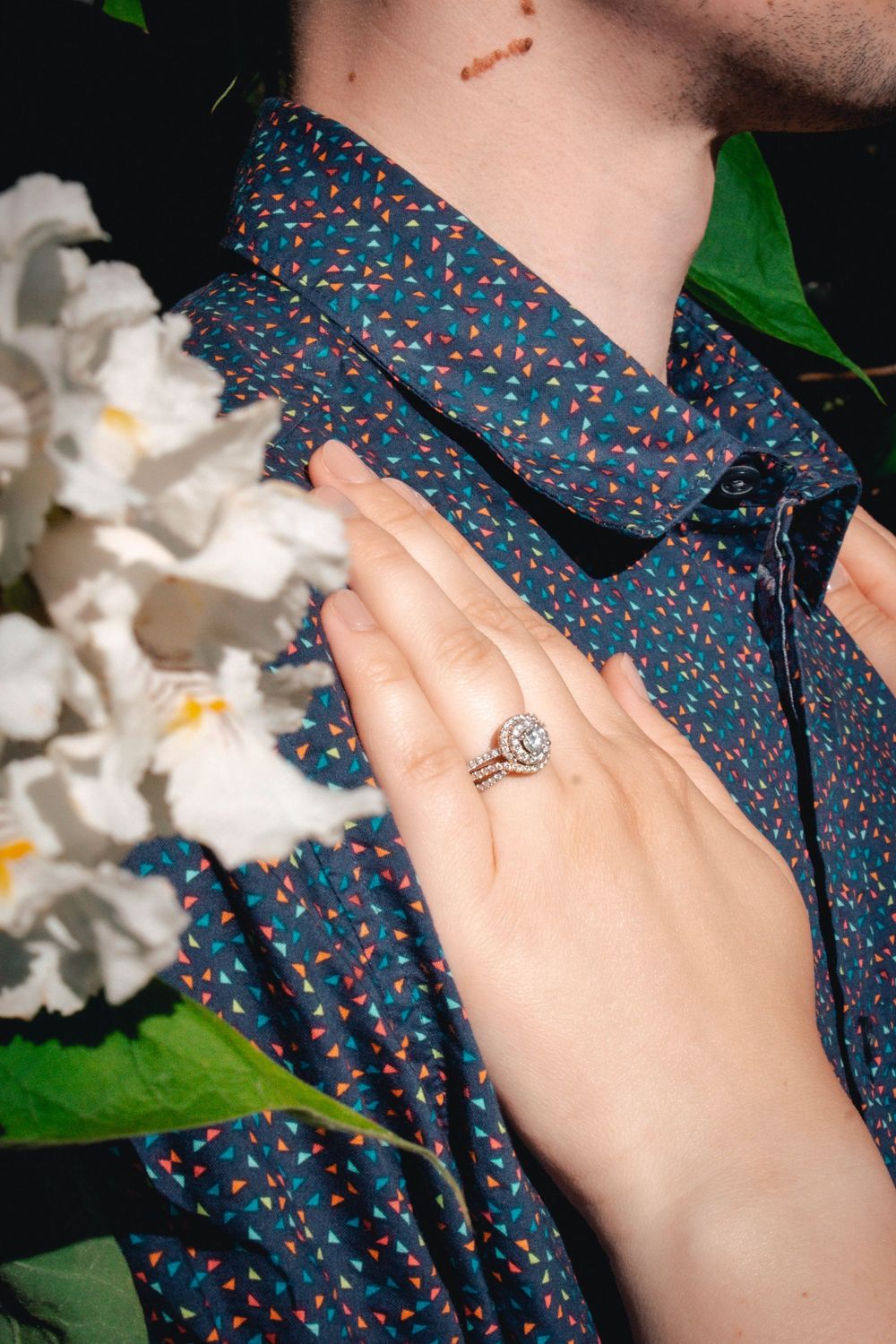 Person wearing a navy blue floral print shirt holds up their hand to show an engagement ring against white flowers.