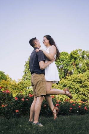 A romantic couple embraces in a garden with blooming roses and green trees on a sunny summer day.