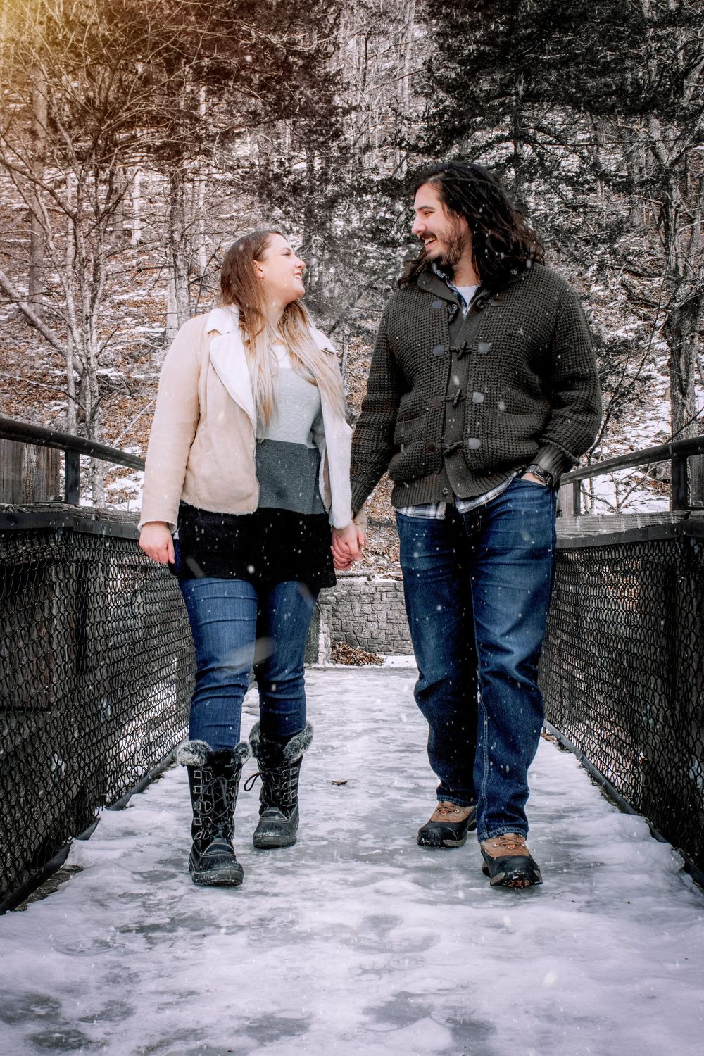 A couple laughs together while walking on a snowy footbridge surrounded by winter trees in a romantic outdoor setting.