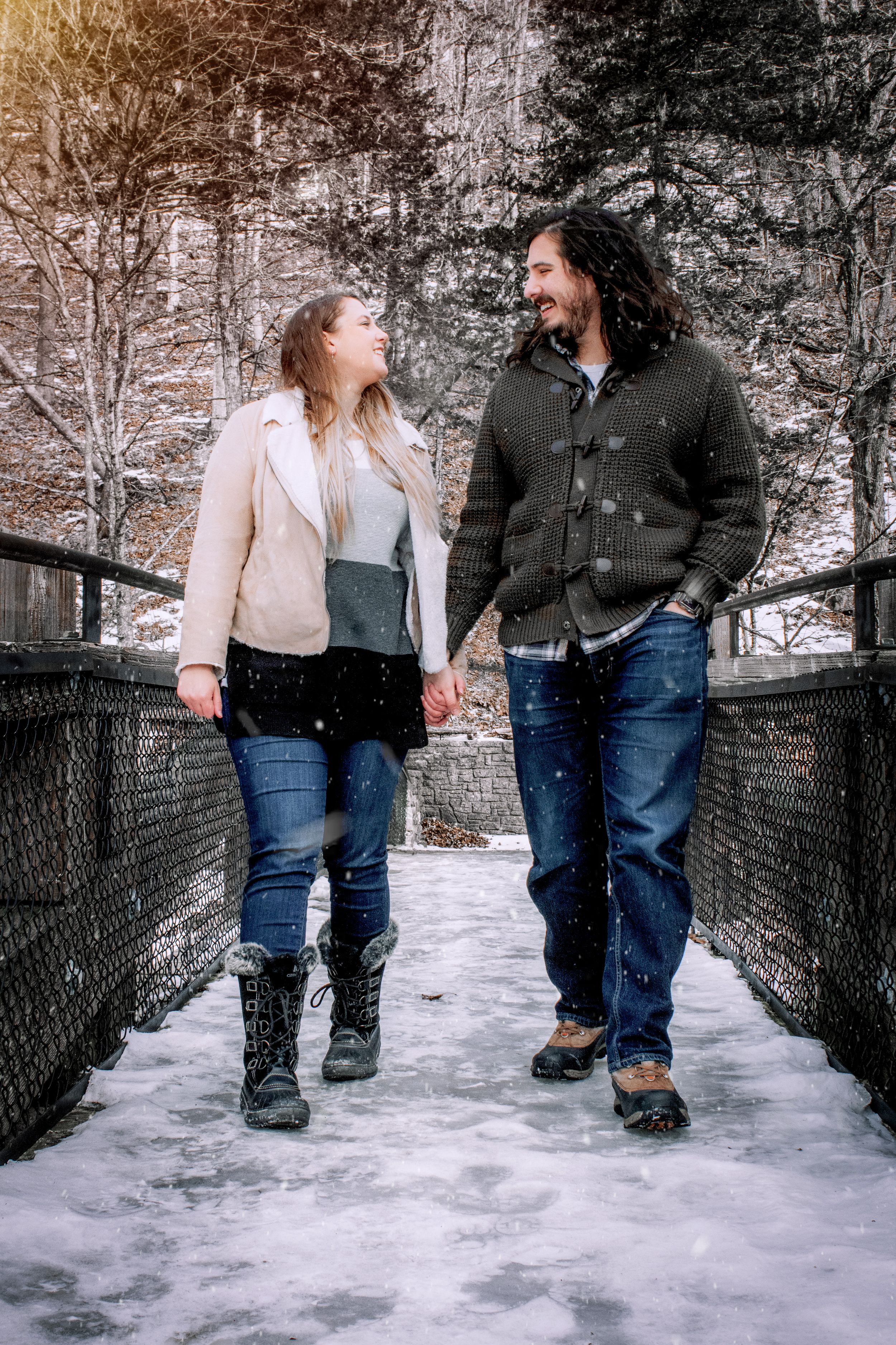 A couple laughs together while walking on a snowy footbridge surrounded by winter trees in a romantic outdoor setting.