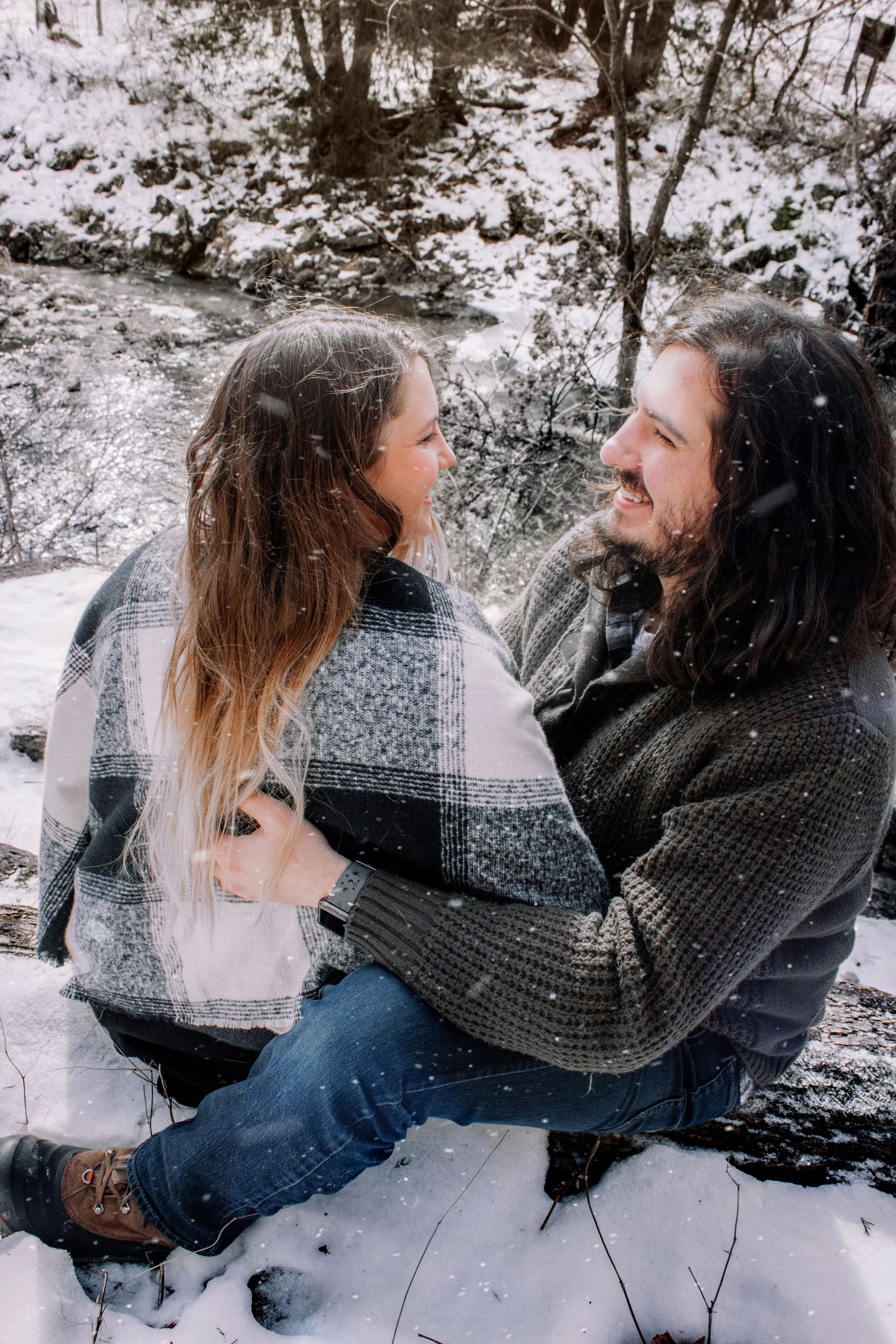 A couple sits together and shares a tender moment in the snow-covered woods wearing cozy winter sweaters.