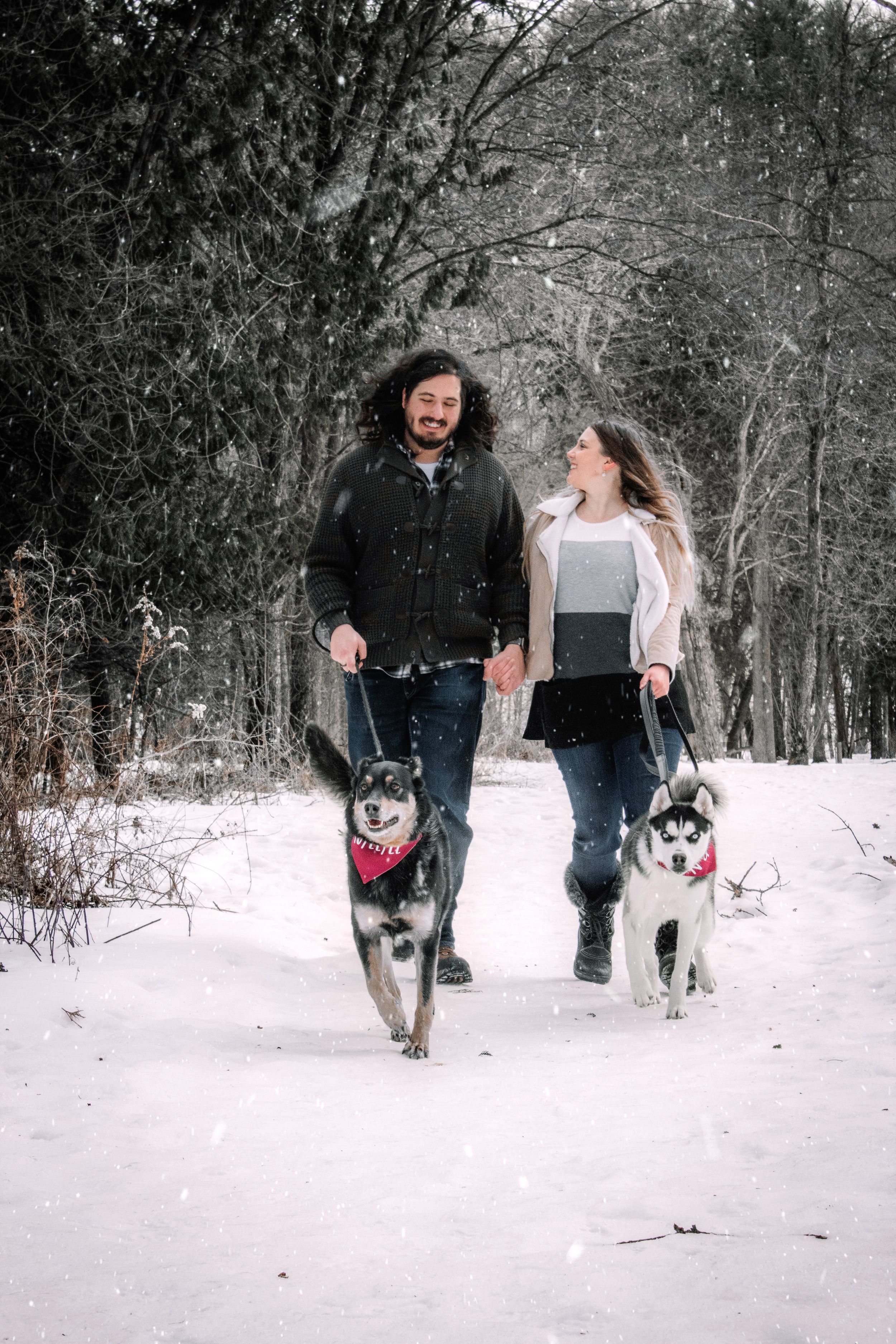 A couple walking two Husky dogs together on a snow-covered path through a winter forest.