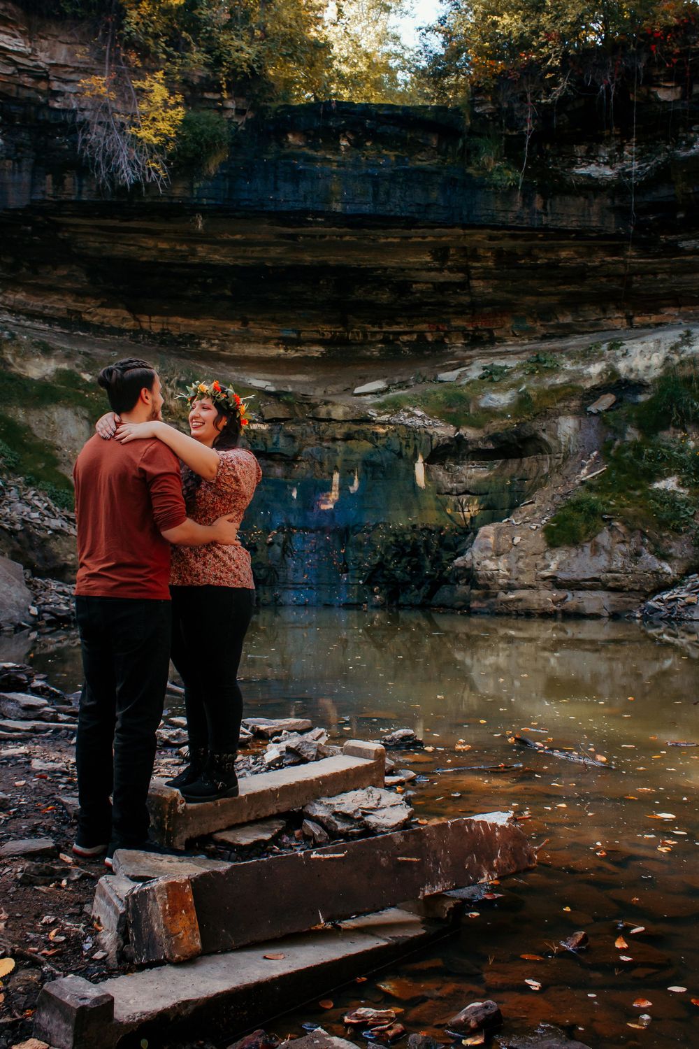 A couple embraces near a scenic waterfall with layered rock formations and autumn leaves scattered around the water pool.