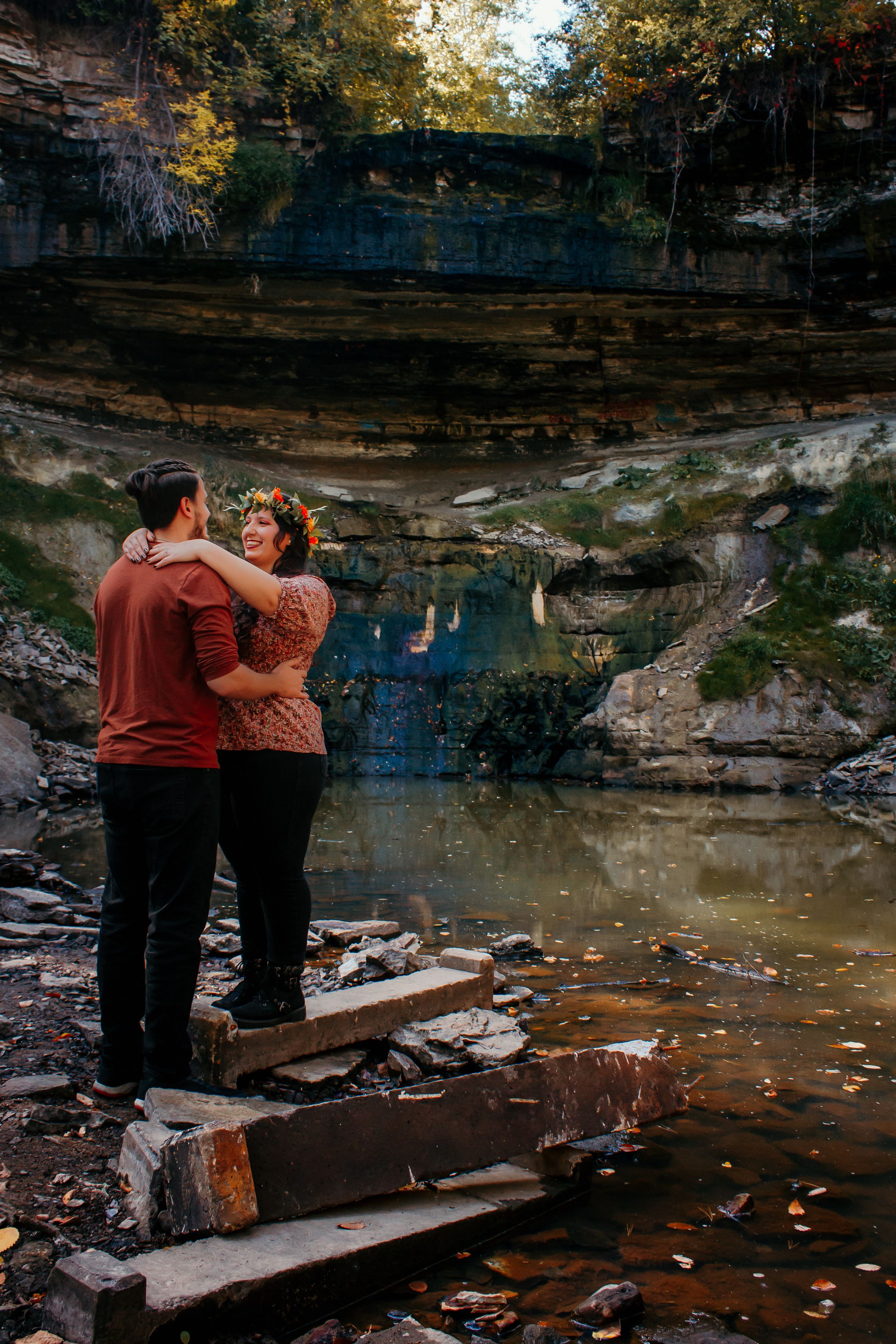 A couple embraces near a scenic waterfall with layered rock formations and autumn leaves scattered around the water pool.