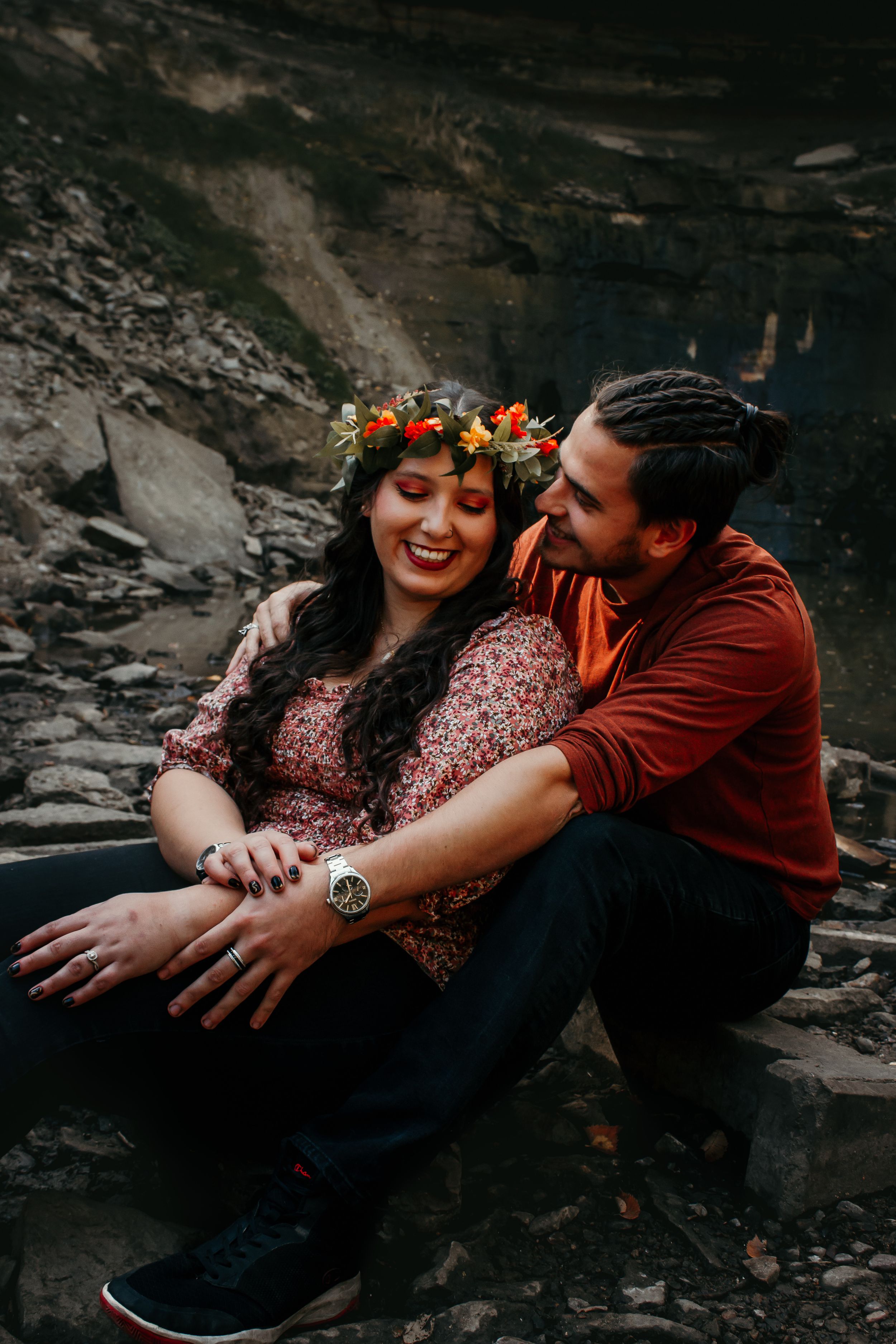 A couple shares an intimate moment sitting on rocky terrain with a floral crown and moody dark background.