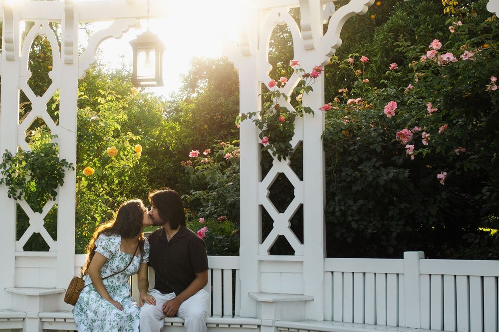 Romantic couple sitting on a white garden gazebo surrounded by blooming roses and greenery at sunset.