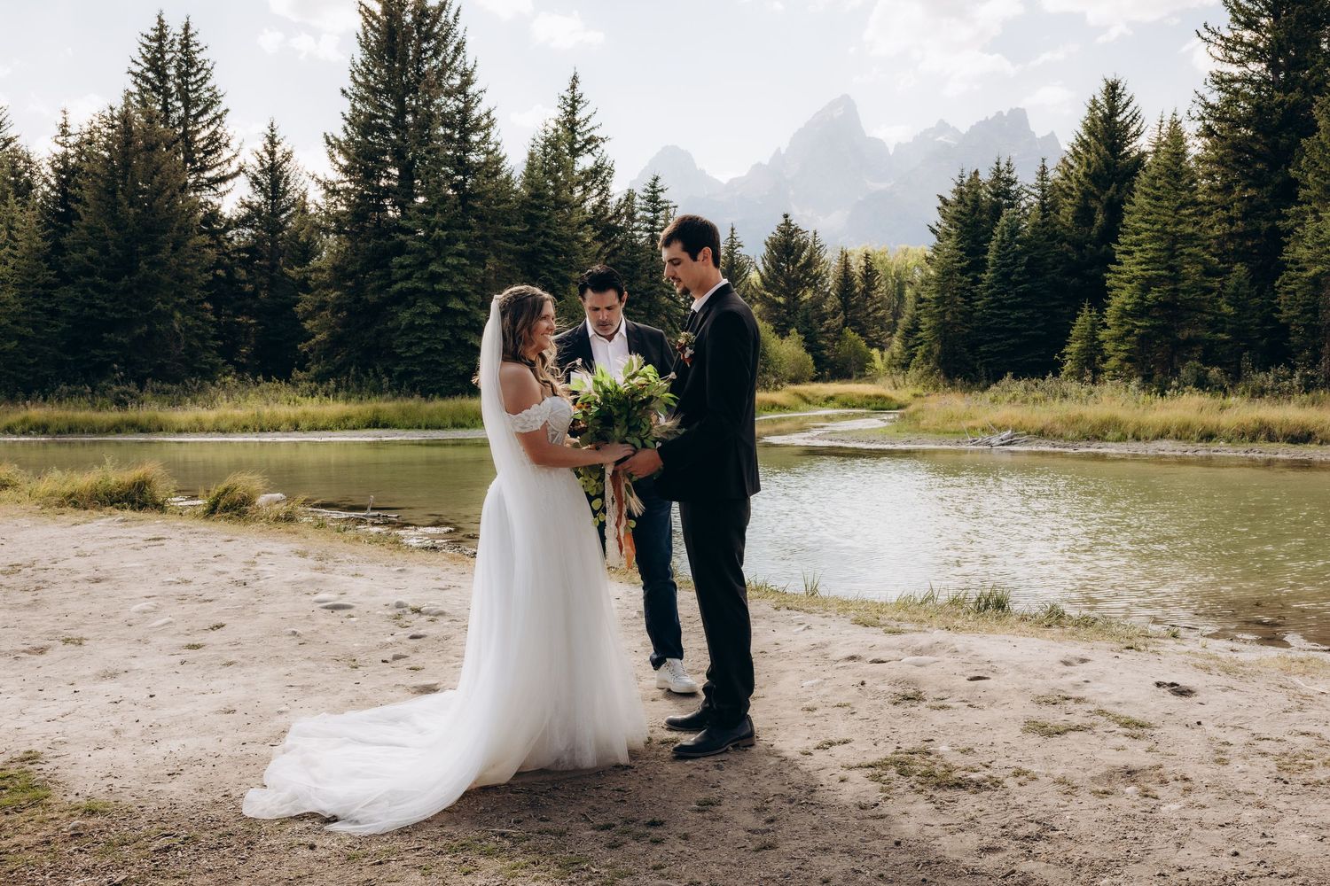A scenic mountain wedding ceremony takes place near evergreen trees and a serene lake in the wilderness.