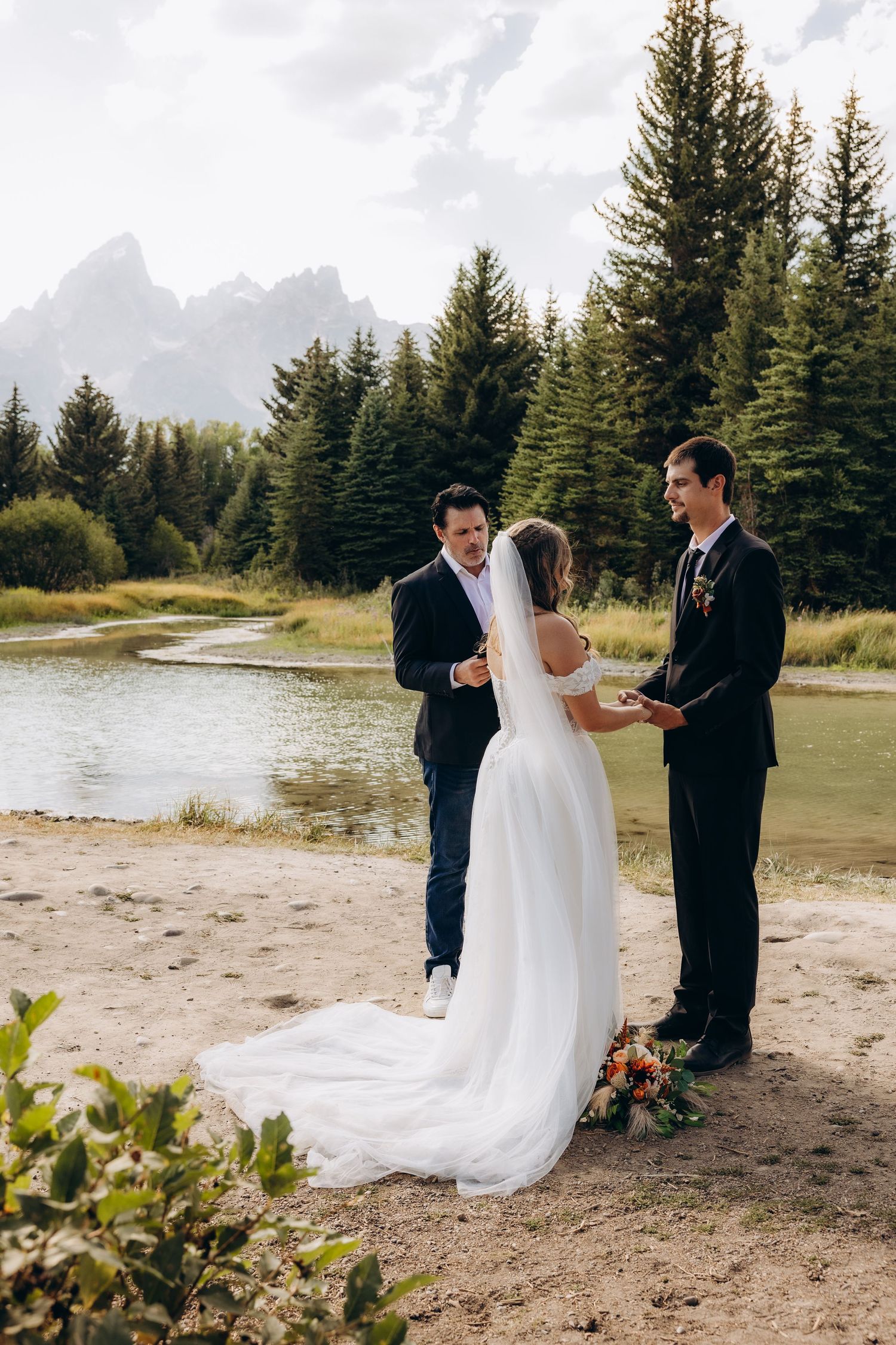 A wedding ceremony takes place on a beach with evergreen trees and mountains in the backdrop.