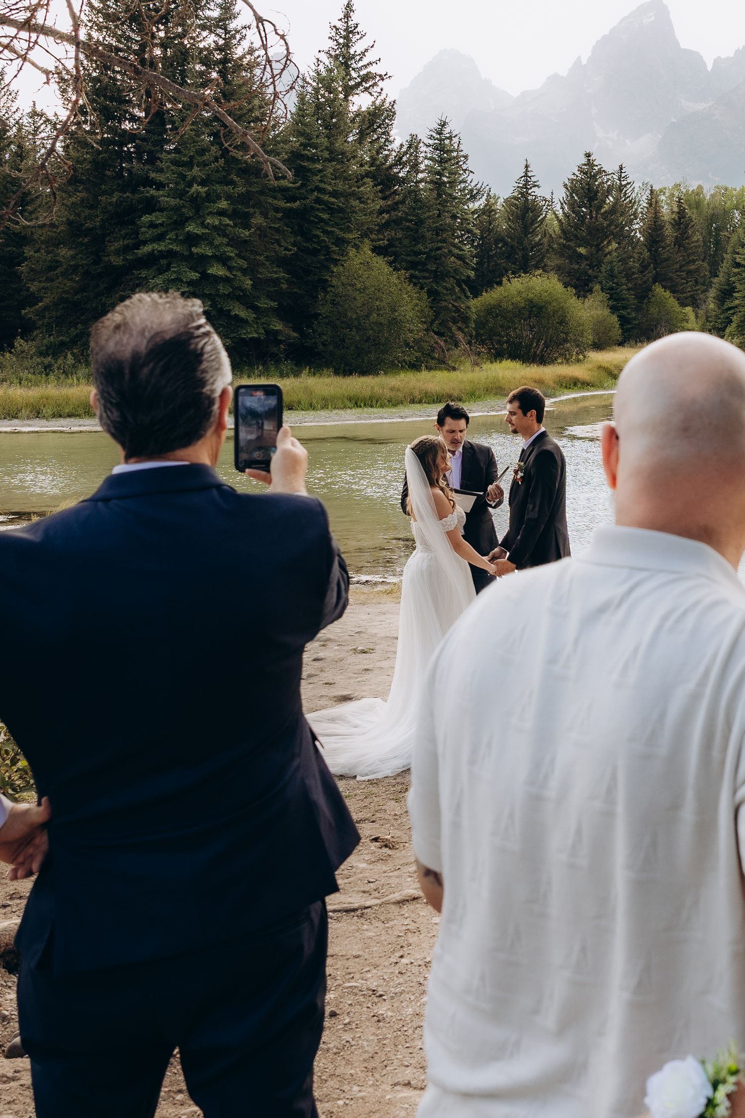 Wedding guests stand at a scenic mountainside ceremony with evergreen trees and peaks in the background.