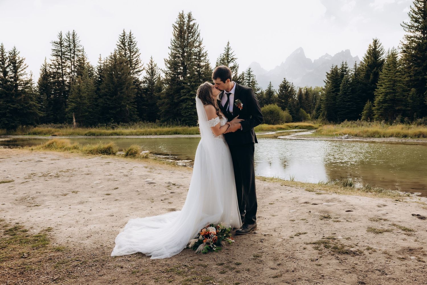 Couple shares intimate wedding moment at mountain lake with evergreen forest and misty peaks in background.