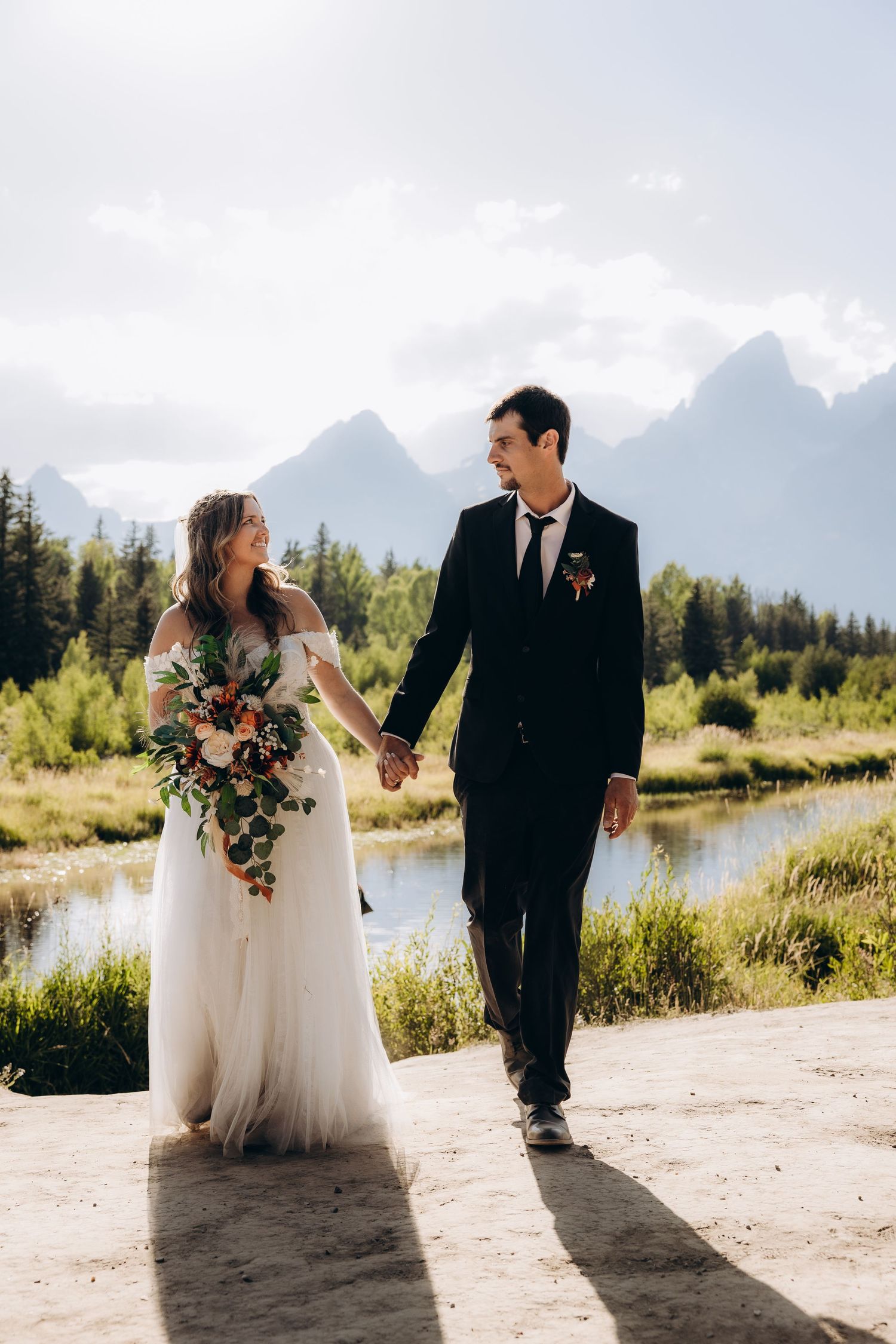 A scenic wedding photoshoot shows a couple posing by a mountain lake with evergreen trees and dramatic peaks in the background.