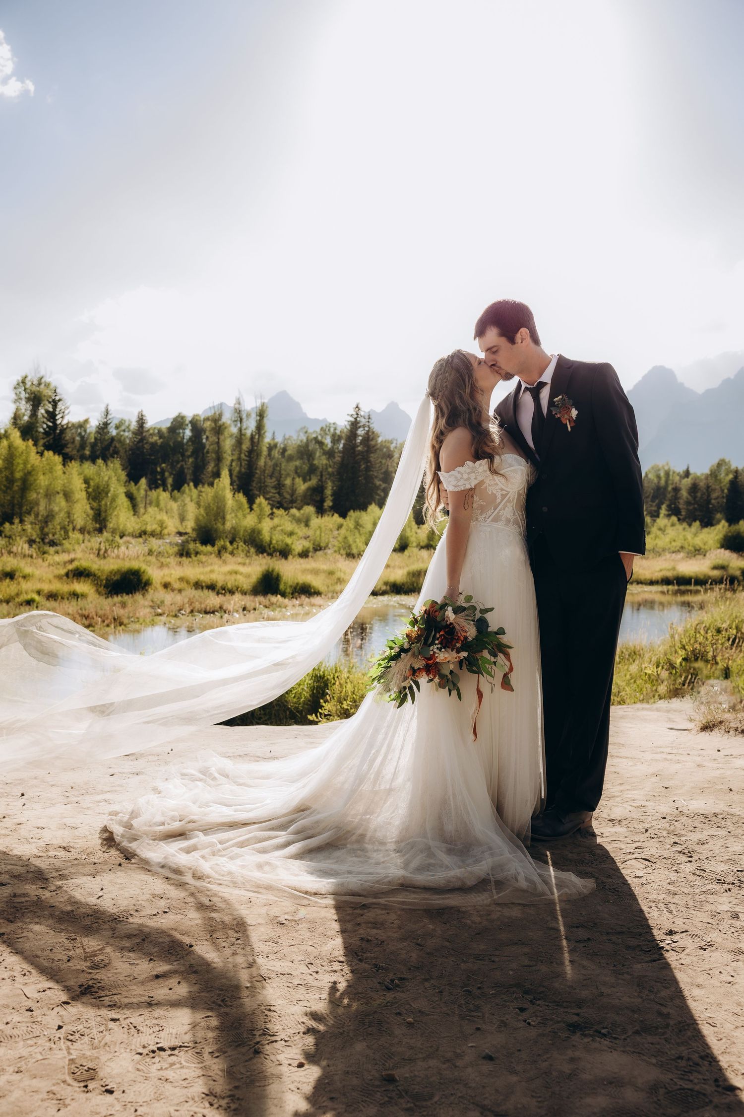 A romantic wedding couple stands against a stunning mountain backdrop as a long veil flows in the wind.