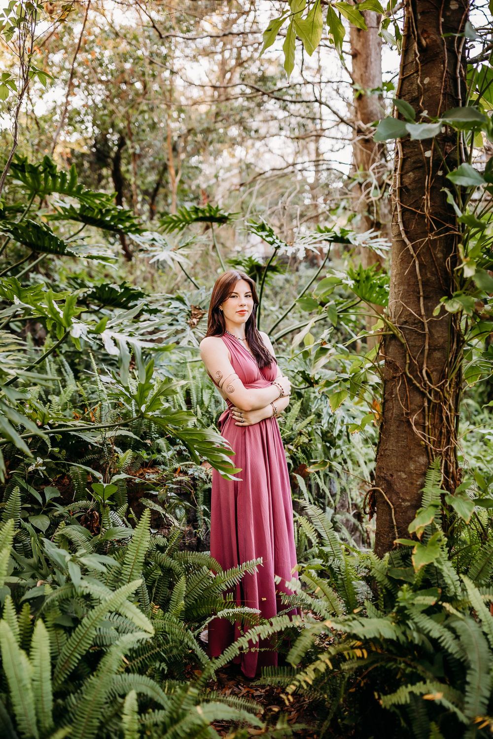 A pink maxi dress contrasts beautifully against lush green ferns and tree trunks in a tranquil forest setting.