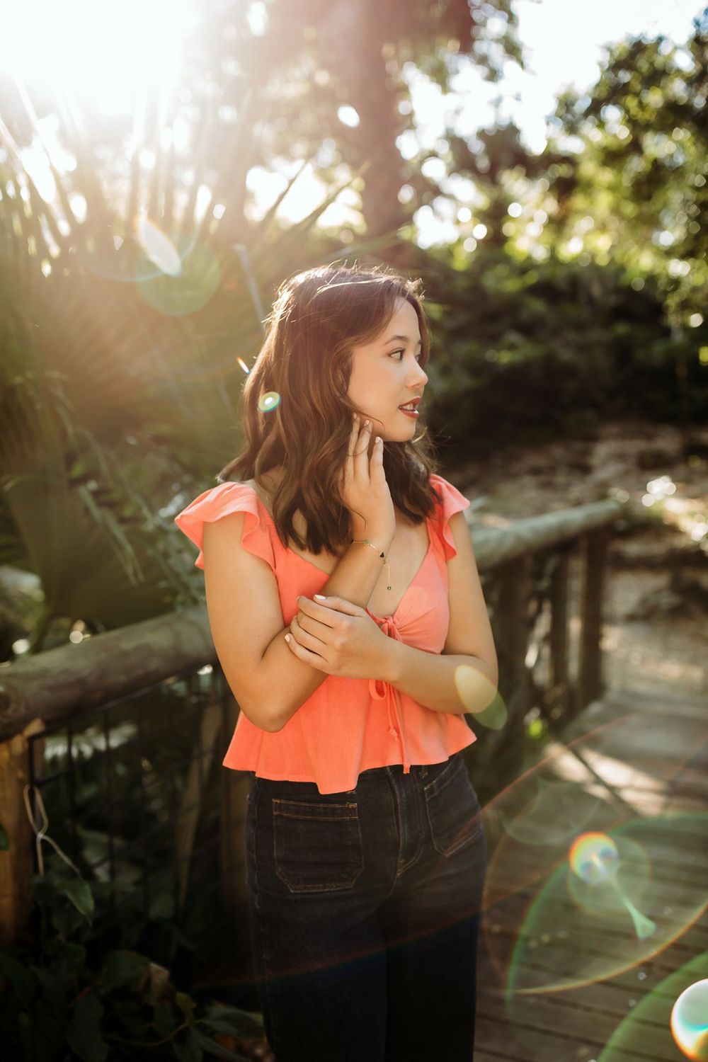 Young woman in coral ruffled top and dark jeans poses outdoors in natural sunlight with lens flare effects.