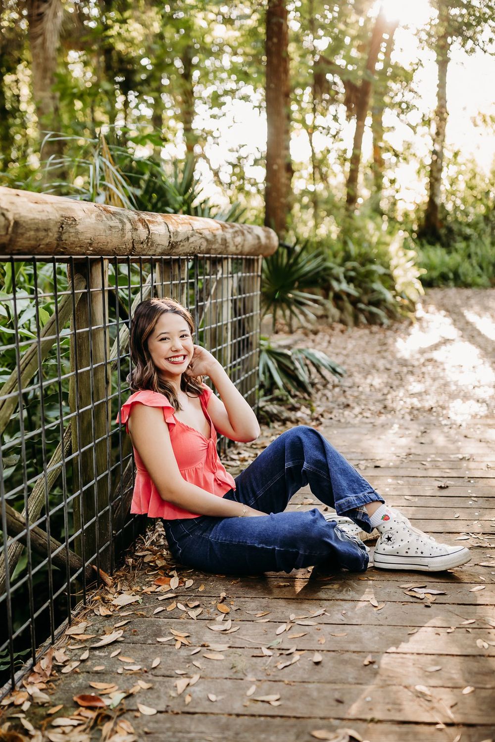 Woman in coral top and jeans relaxing on wooden boardwalk in dappled sunlight through forest trees.