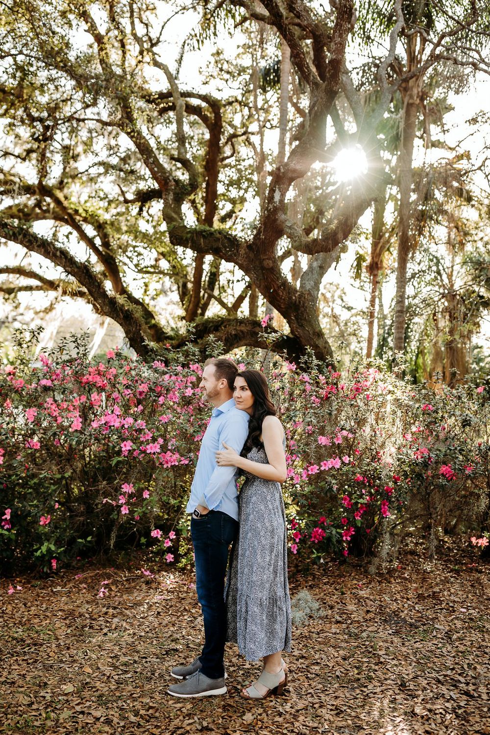 A couple shares a tender moment in a garden filled with pink blooming azalea bushes and oak trees at sunset.