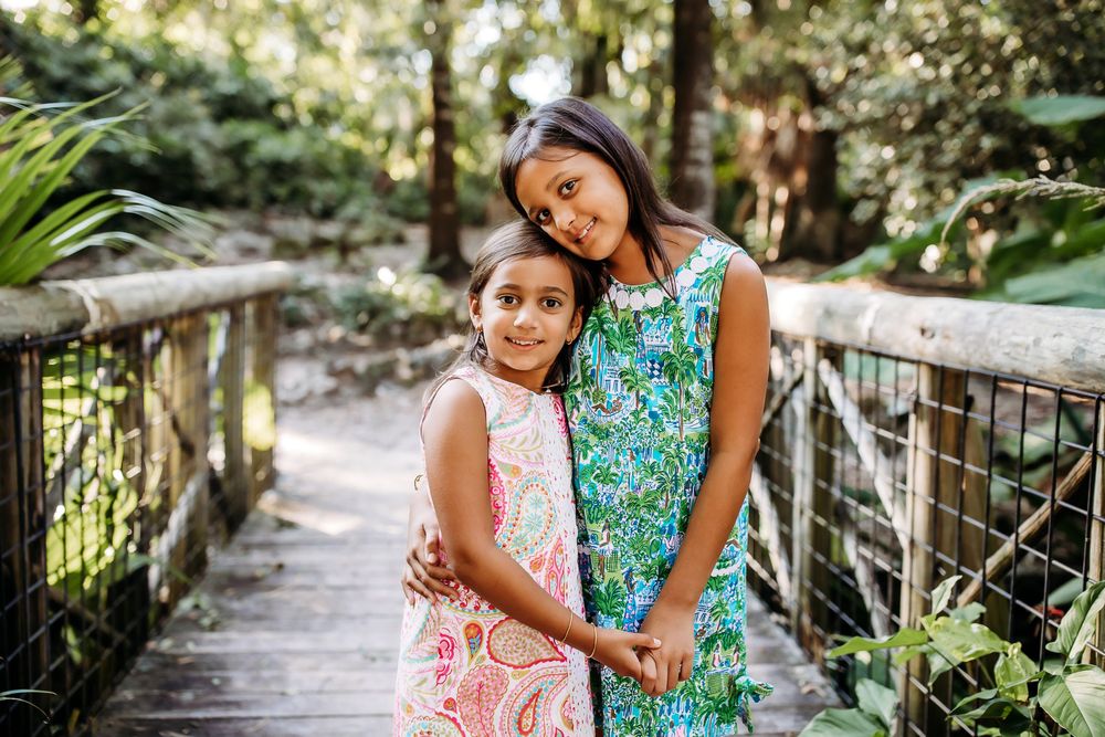 Two people wearing colorful dresses share an affectionate moment on a wooden bridge in a lush garden setting.