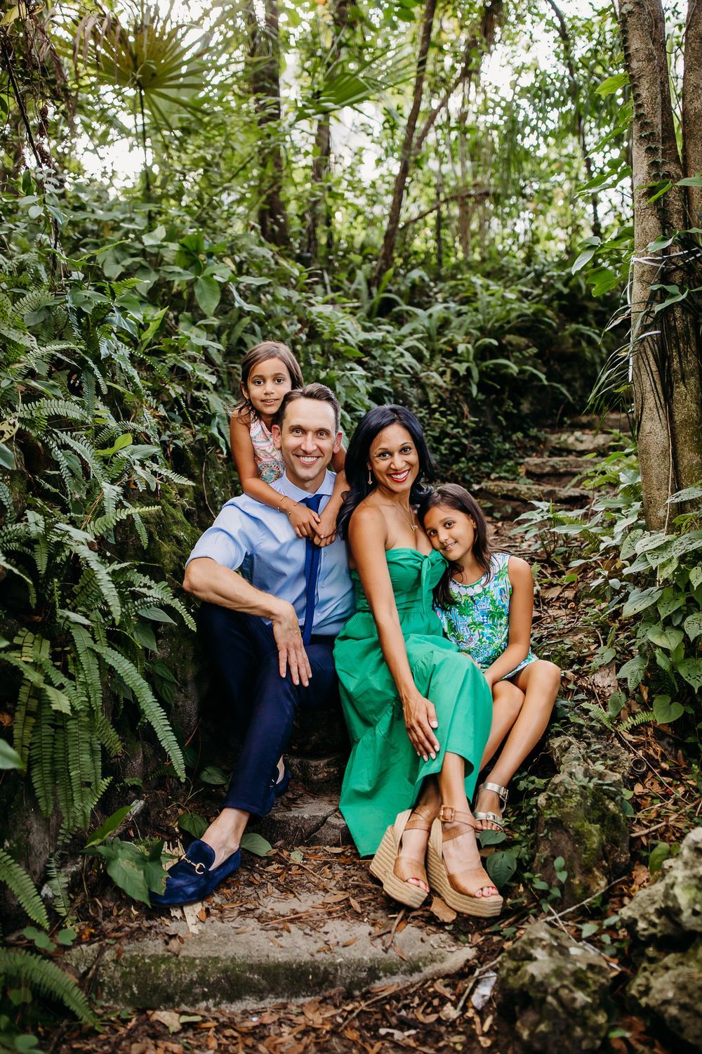 A family sits together on outdoor stone steps surrounded by lush tropical greenery and foliage in matching green outfits.