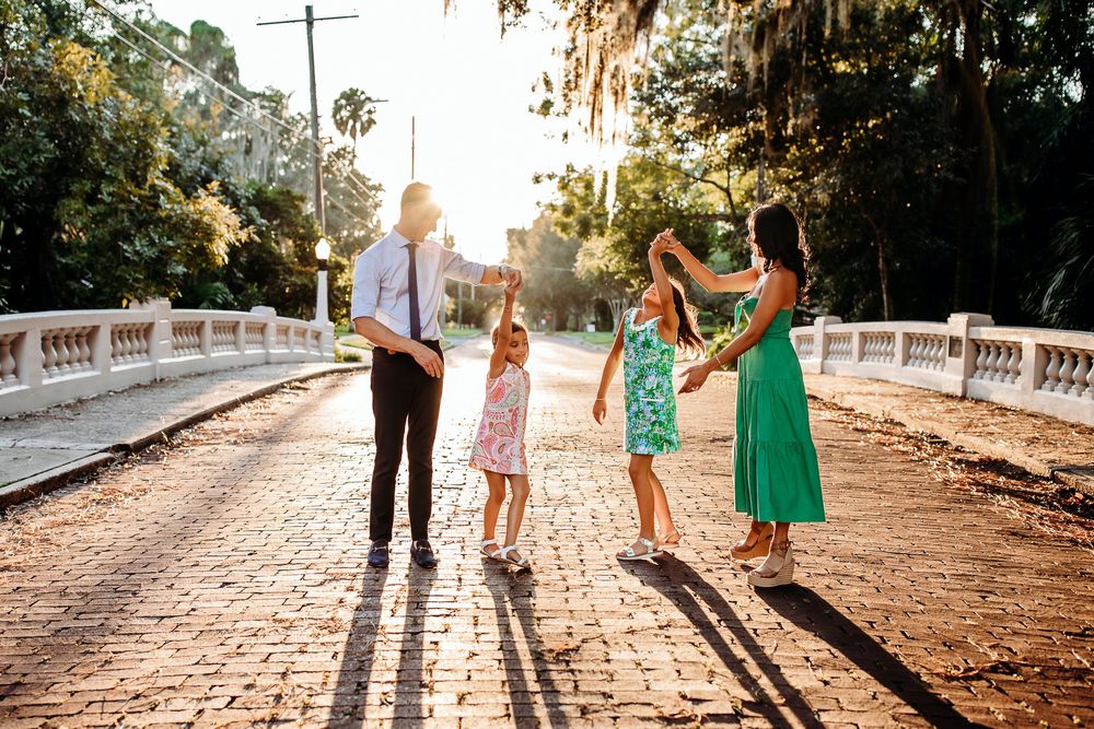 A family dances together on a sunlit cobblestone bridge during golden hour wearing coordinated summer outfits.