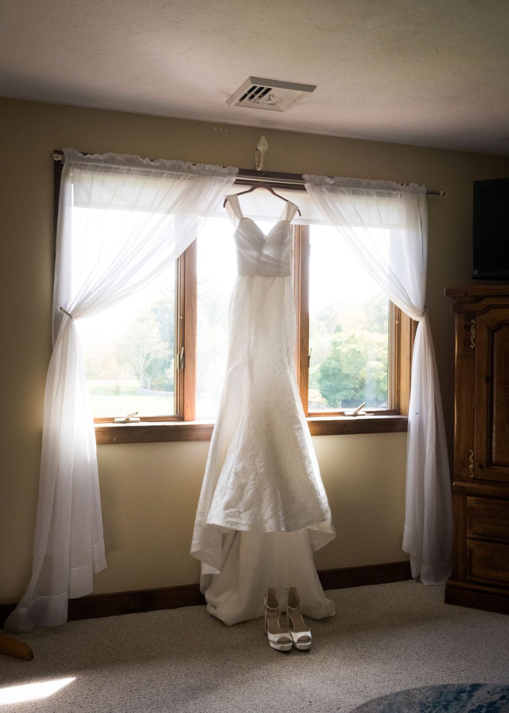A white wedding dress hangs elegantly in a sunlit room with sheer curtains and white high heel shoes visible on the windowsill.