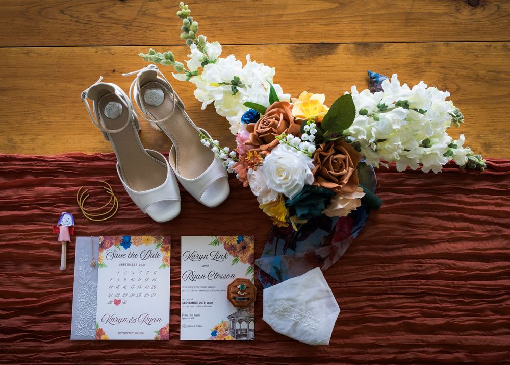 Wedding details flatlay with white shoes, floral bouquet and invitation cards on wooden and burgundy textured surfaces.