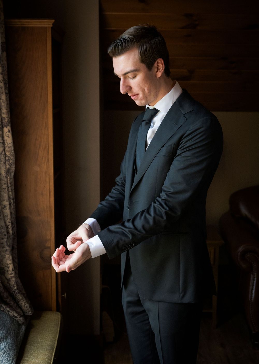 A person in a dark suit checks their watch while standing in a dimly lit room with wooden columns.