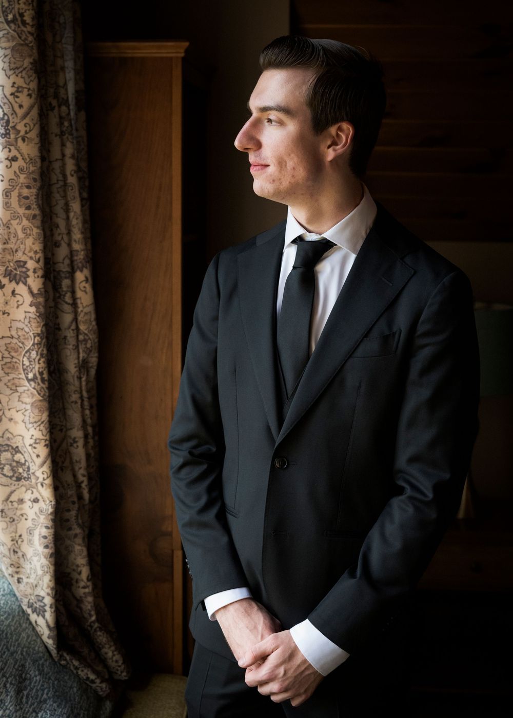 A suited figure gazes thoughtfully to the side near an elegant curtained window.