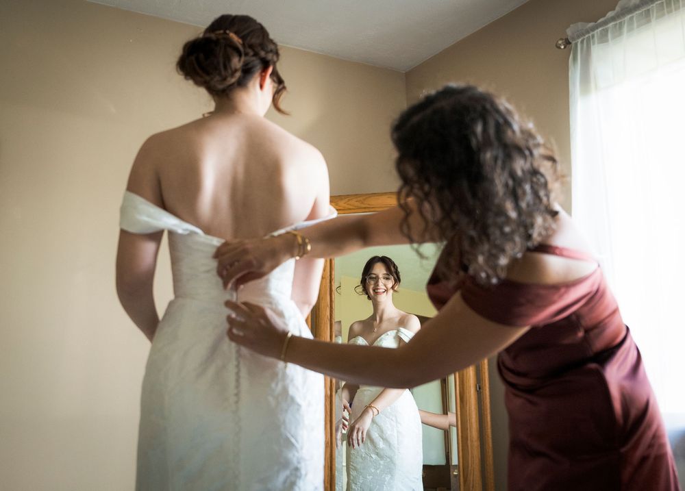 A person in a white wedding dress gets help with final adjustments while looking in a mirror during wedding preparations.