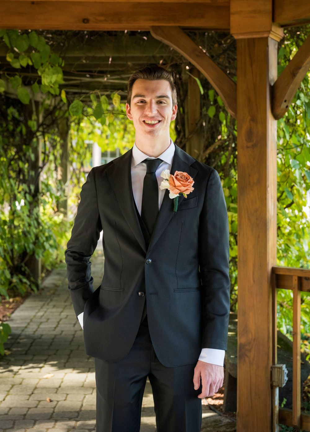 A suited figure with a coral boutonniere stands smiling under a vine-covered garden pergola.