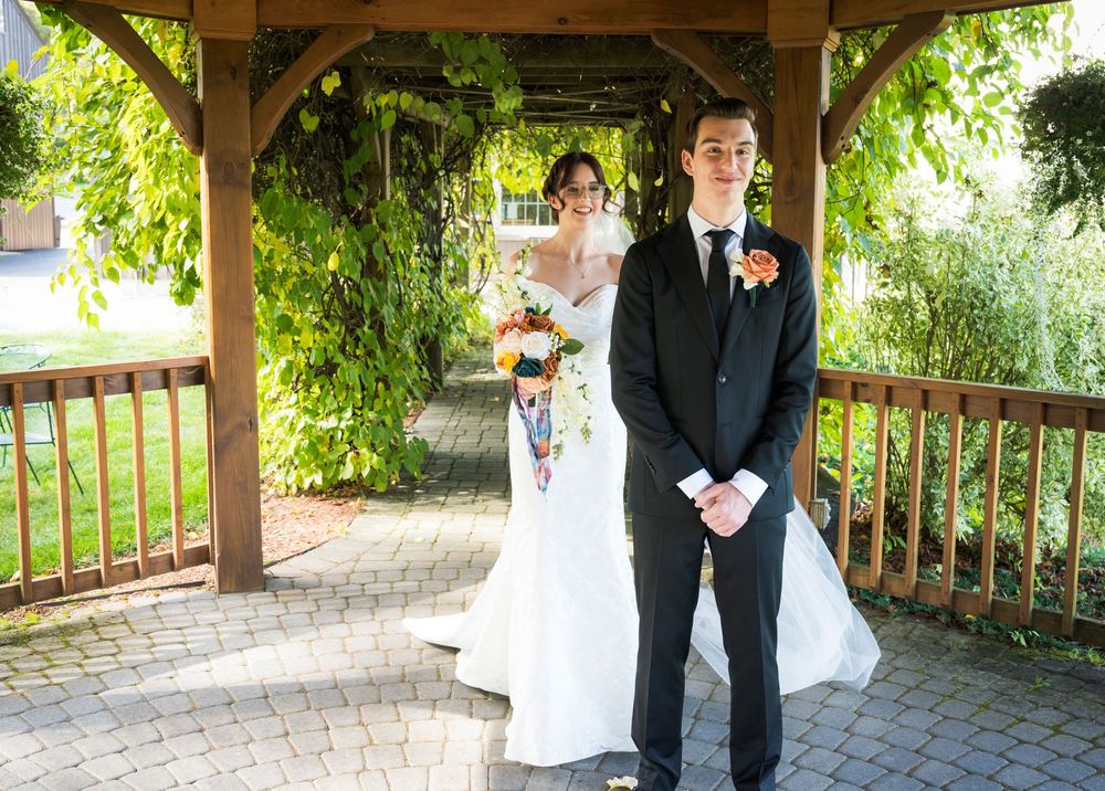 Wedding couple stands under a wooden gazebo surrounded by lush greenery and flowers.