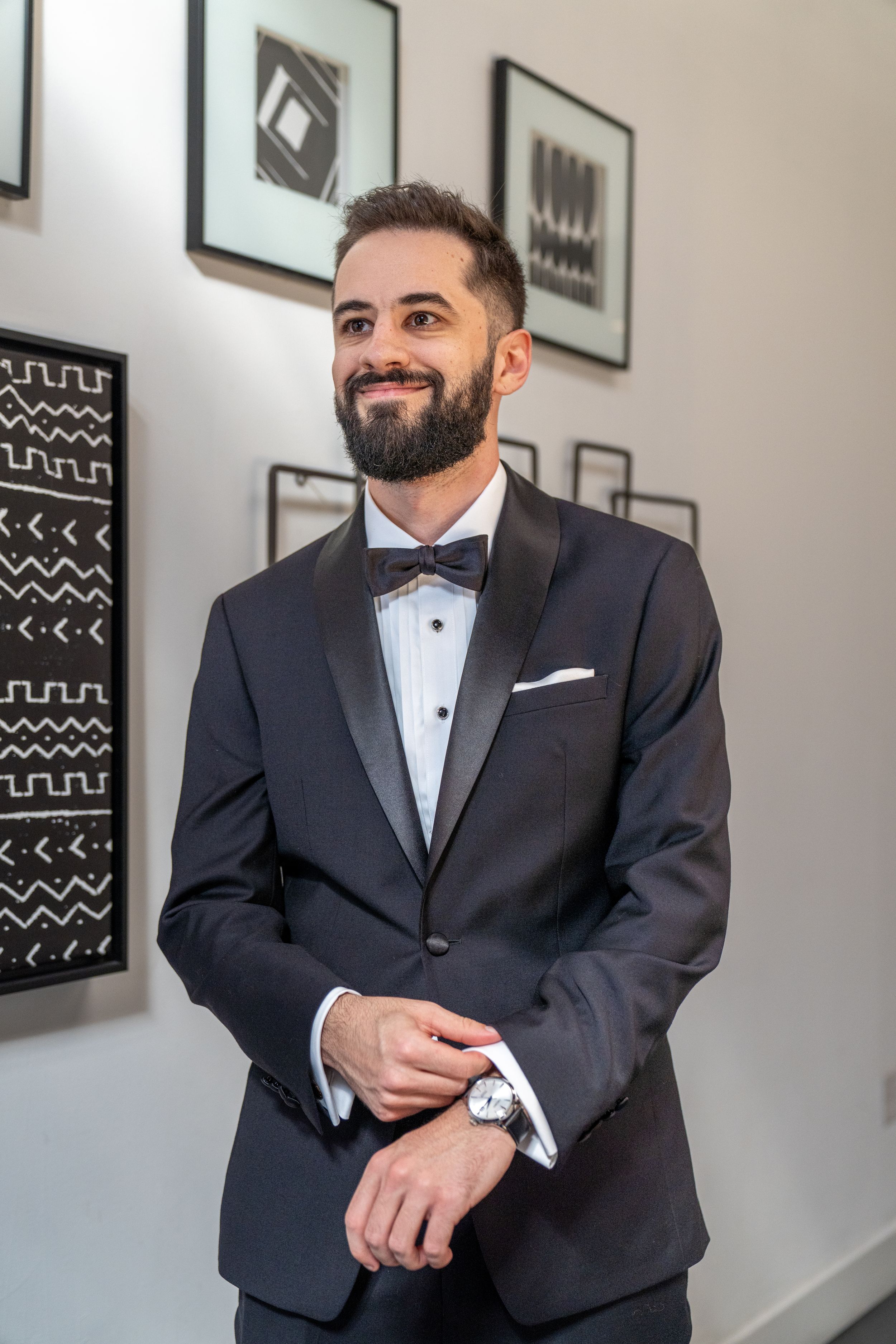 Smiling bearded man in black tuxedo with bow tie and watch, posing indoors near framed wall art.