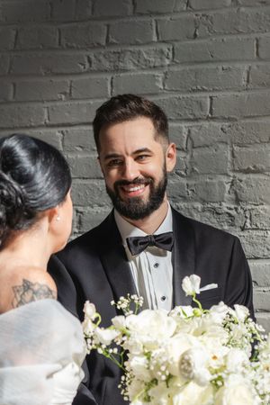 Smiling bearded groom in black tuxedo looking at bride holding white floral bouquet against gray brick wall.