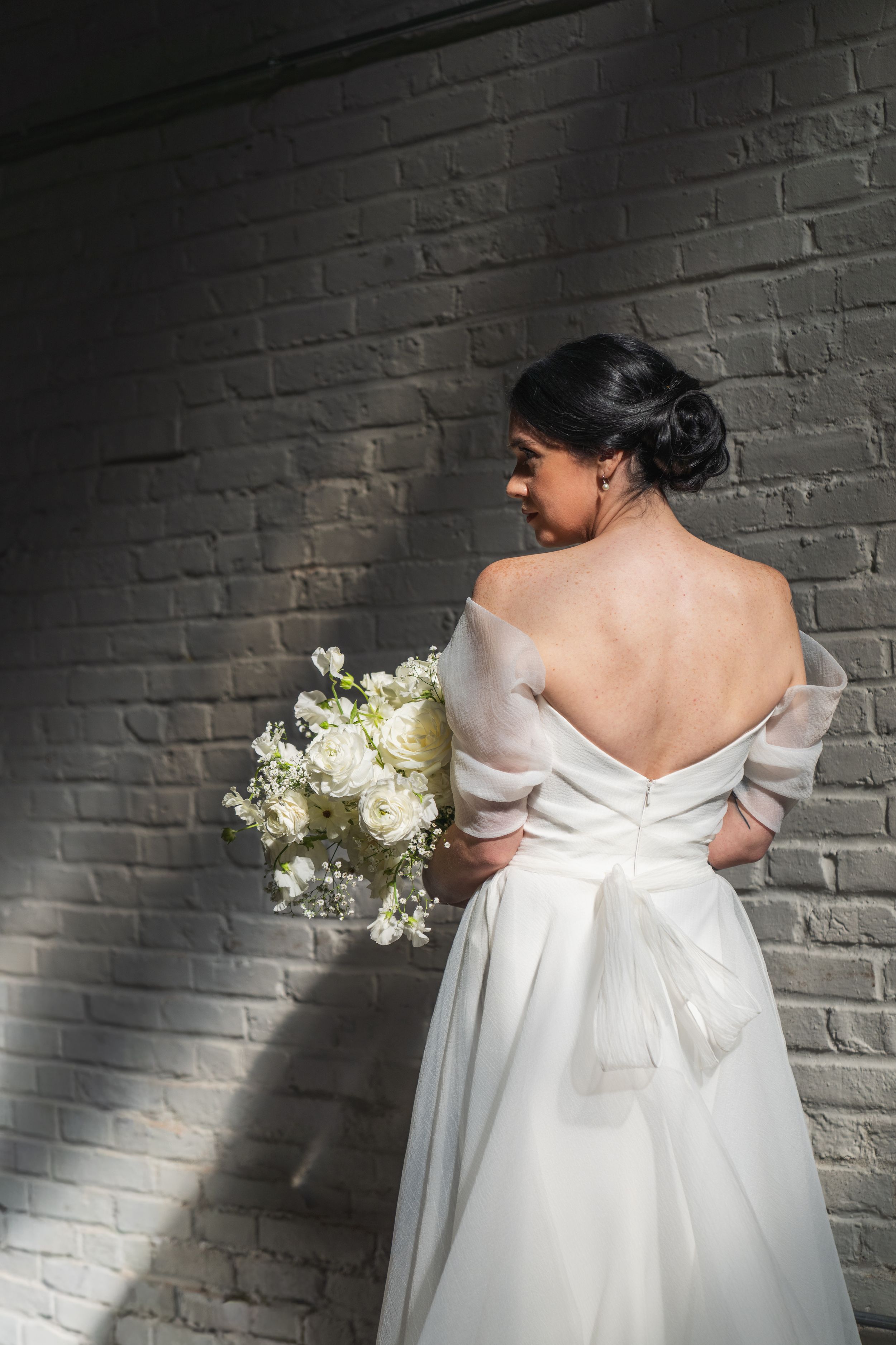 Bride in backless white gown with bow holds floral bouquet against gray brick wall.